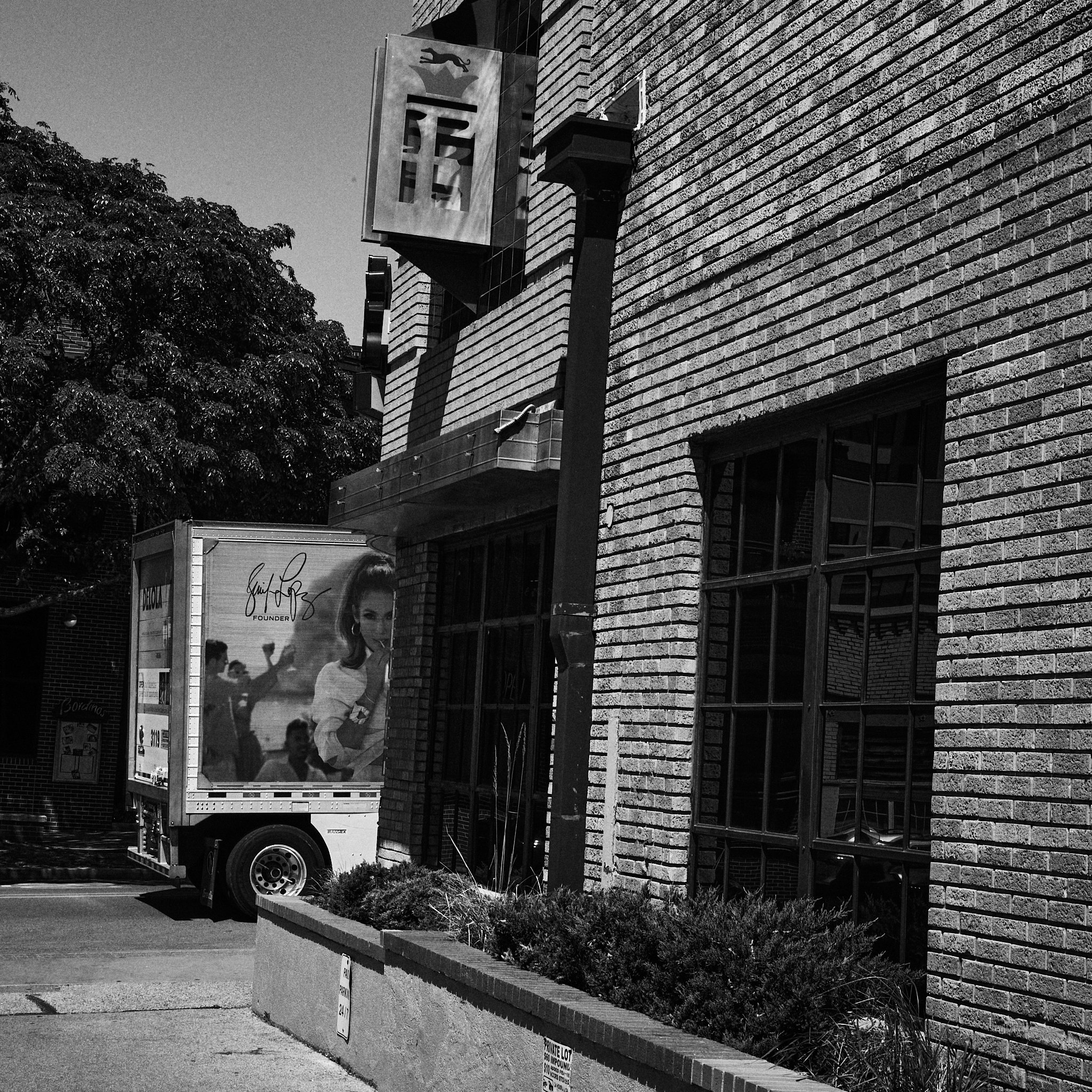 Black and white photo of a brick building with a street sign. A truck with an advertisement featuring a woman holding a phone is parked nearby. There is a large sign with a stylized logo on the building.