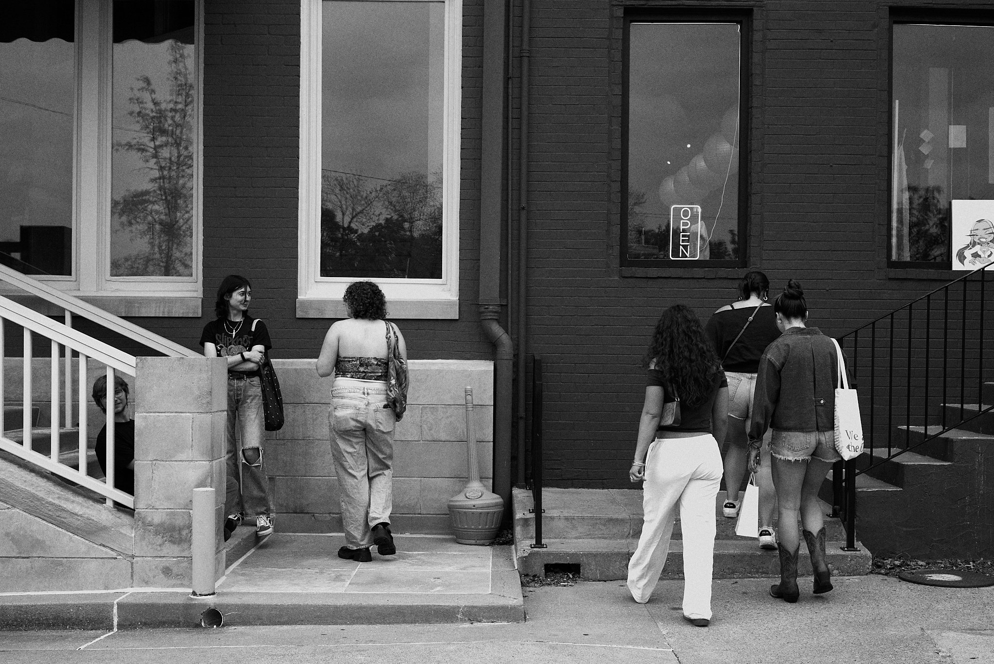 A group of seven young women standing outside a building, some walking up stairs, some talking, with a sign reading 'OPEN' in the window.
