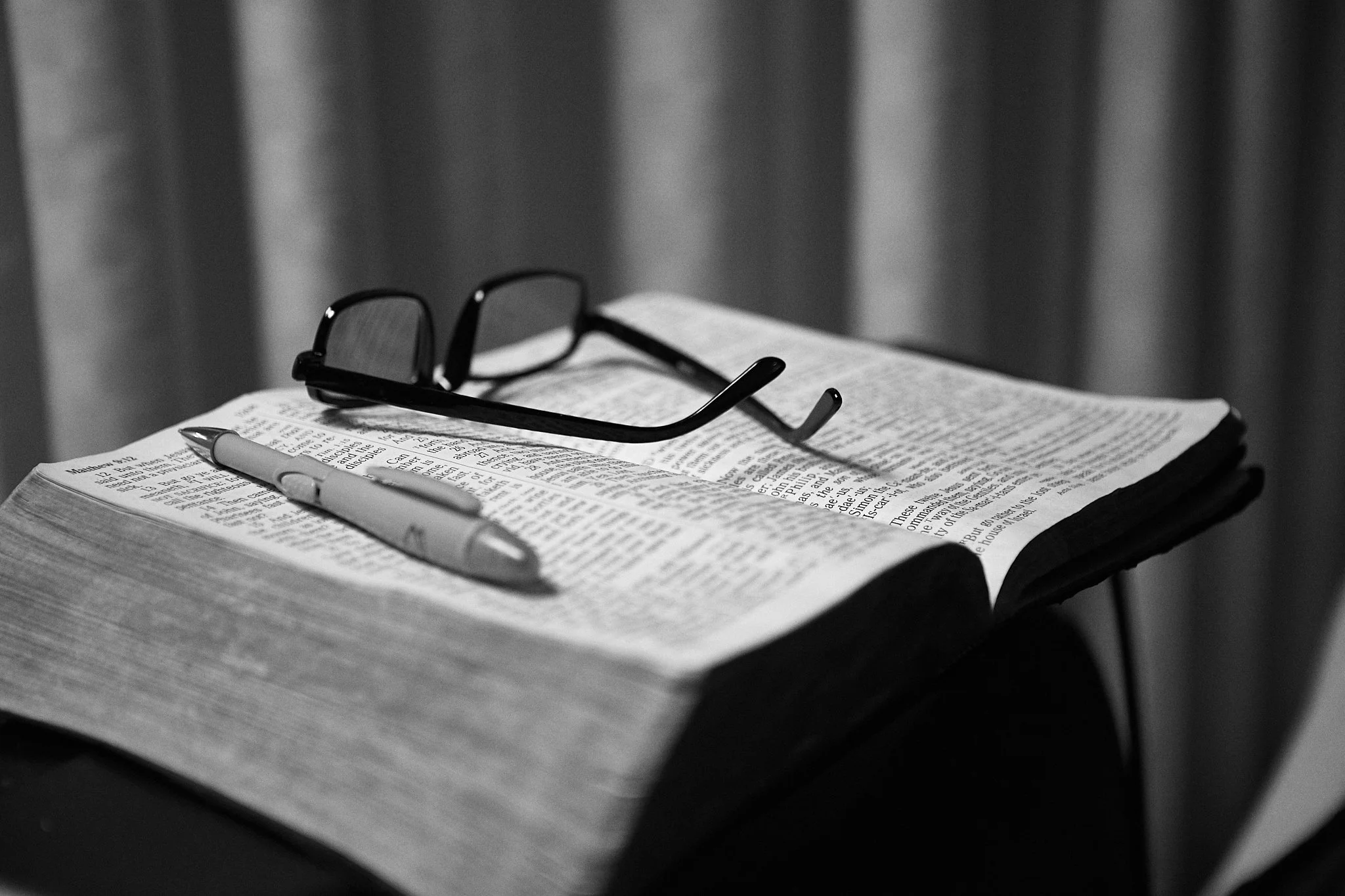 A black and white photograph of reading glasses, a pen, and an open Bible on a table or desk, with curtains in the background.