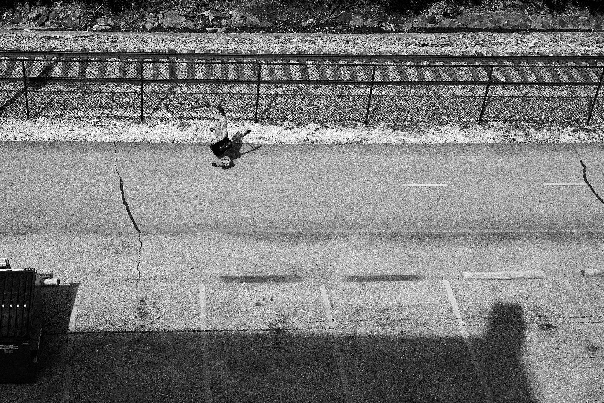 A person walking down a street carrying a guitar, viewed from above in black and white.