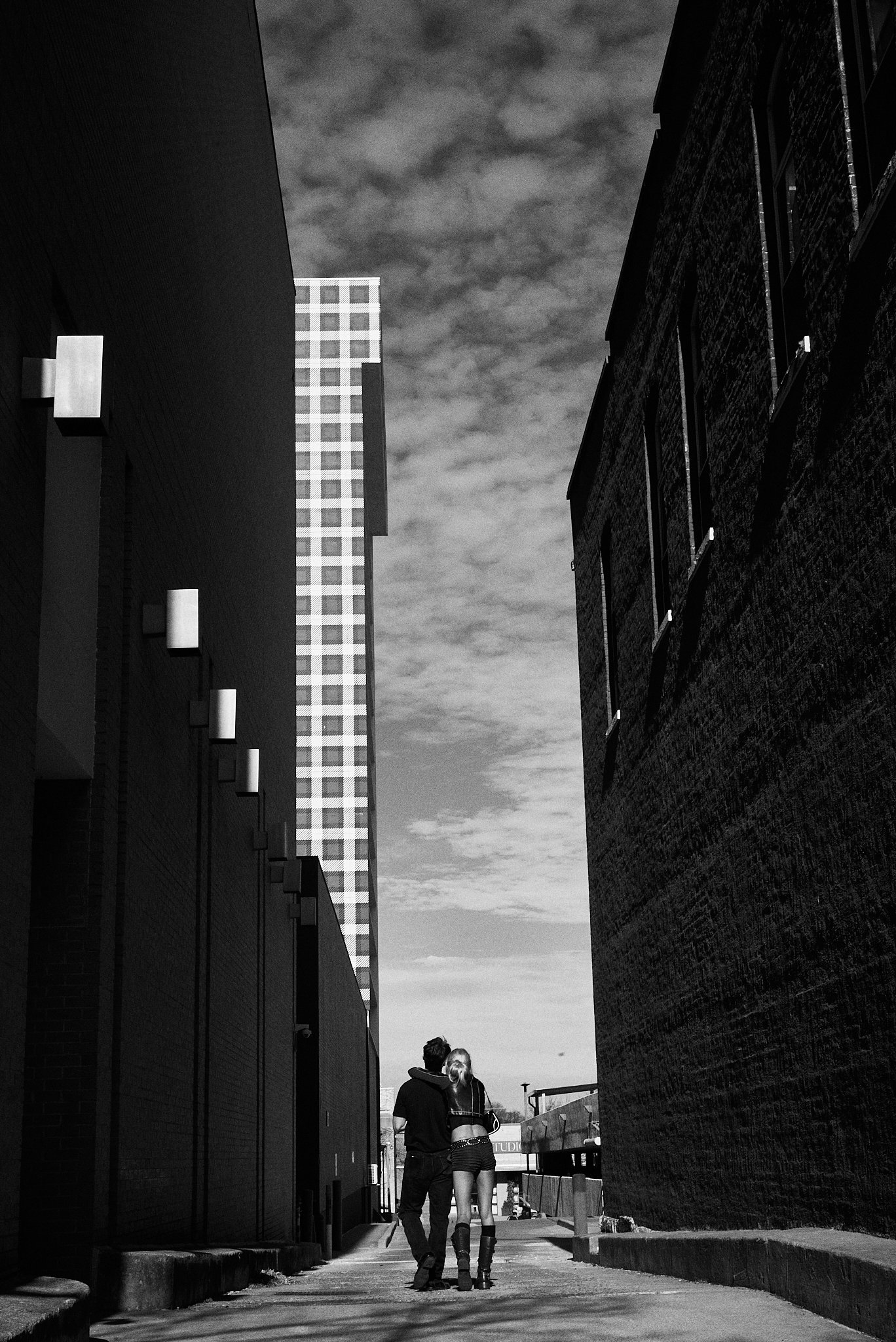Two people, a man and a woman, walking together in a narrow alleyway between tall buildings on a partly cloudy day.