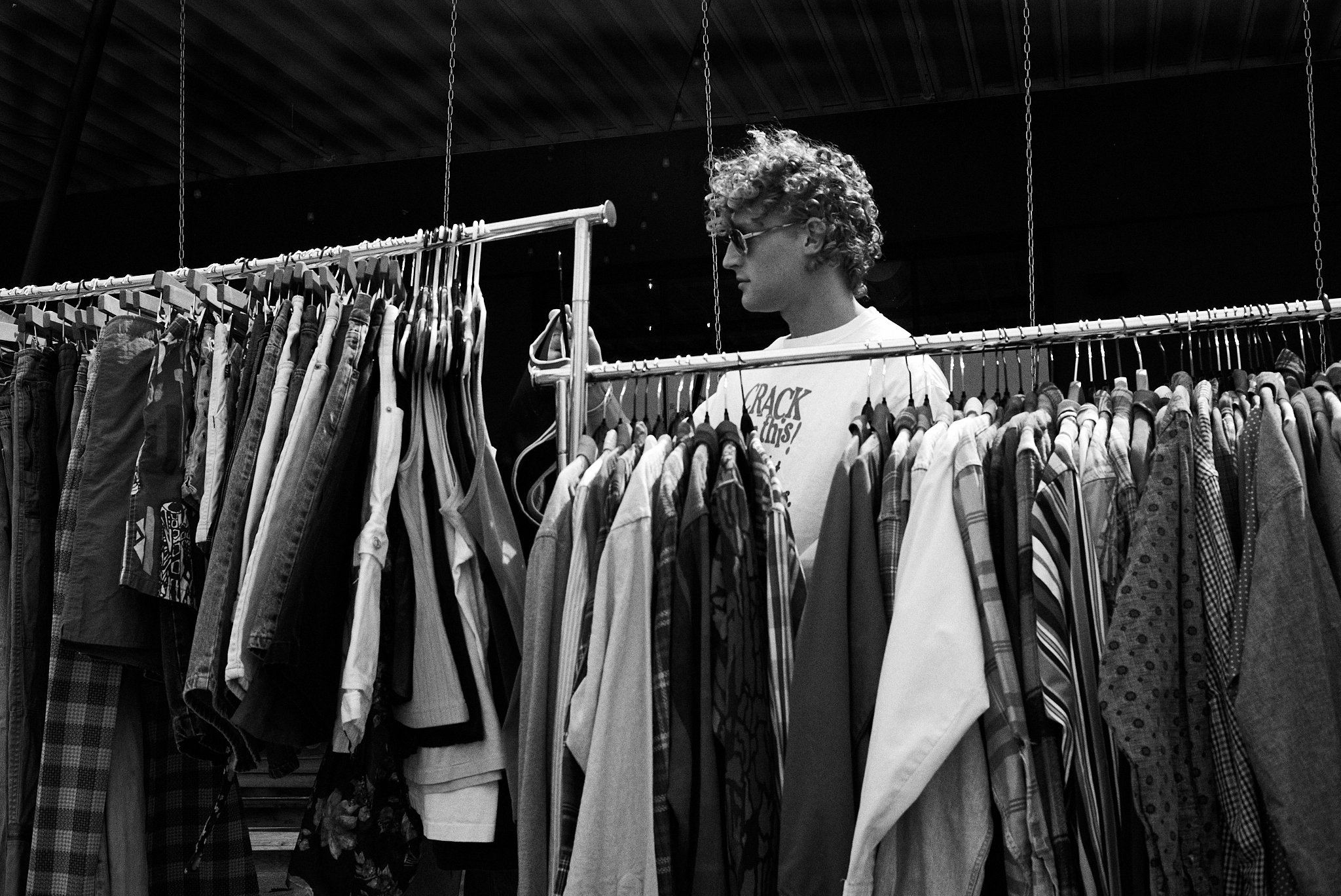 Person with curly hair and glasses browsing clothes on rack in a store.