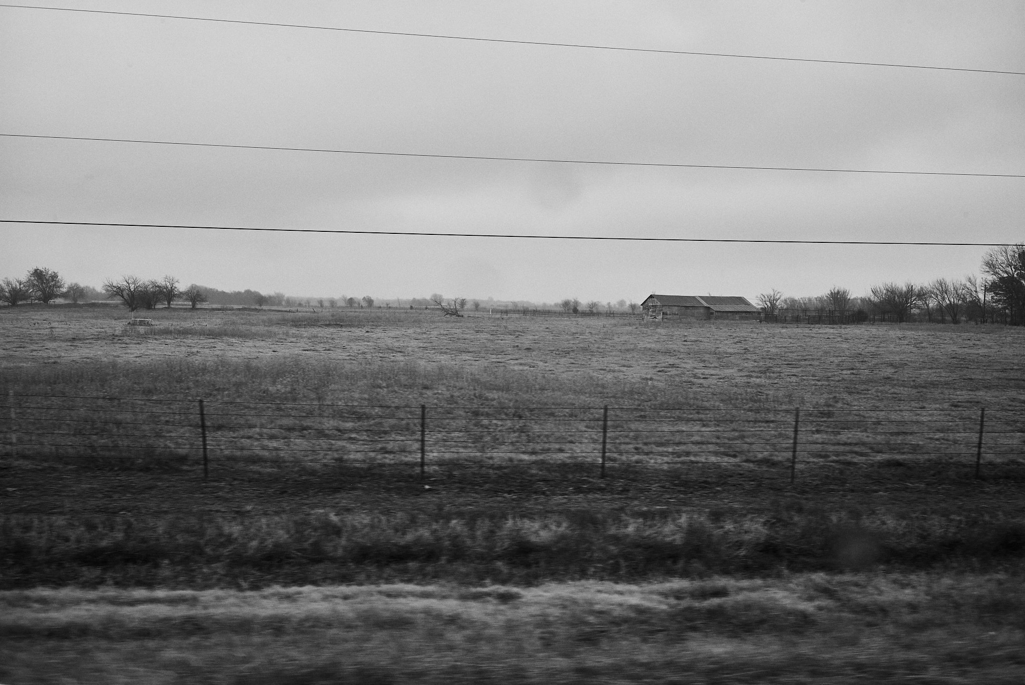 Black and white photo of an open field with a fence in the foreground, a barn in the distance, and a few trees scattered across the landscape under a cloudy sky.