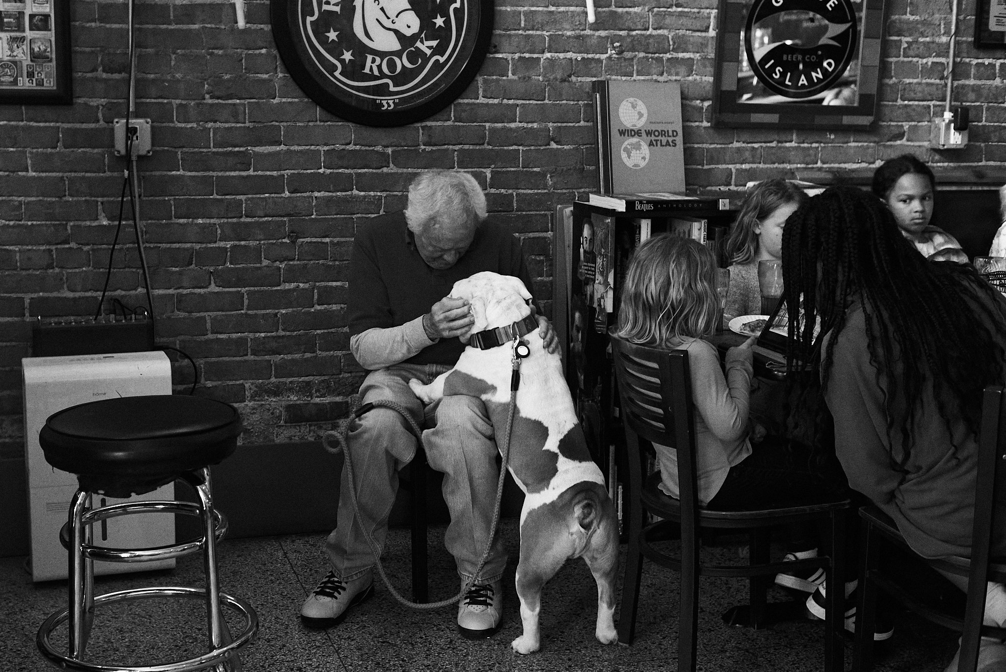 An elderly man sitting at a table in a restaurant with a large dog with a white coat and brown spots, sitting on the floor, leaning against him.