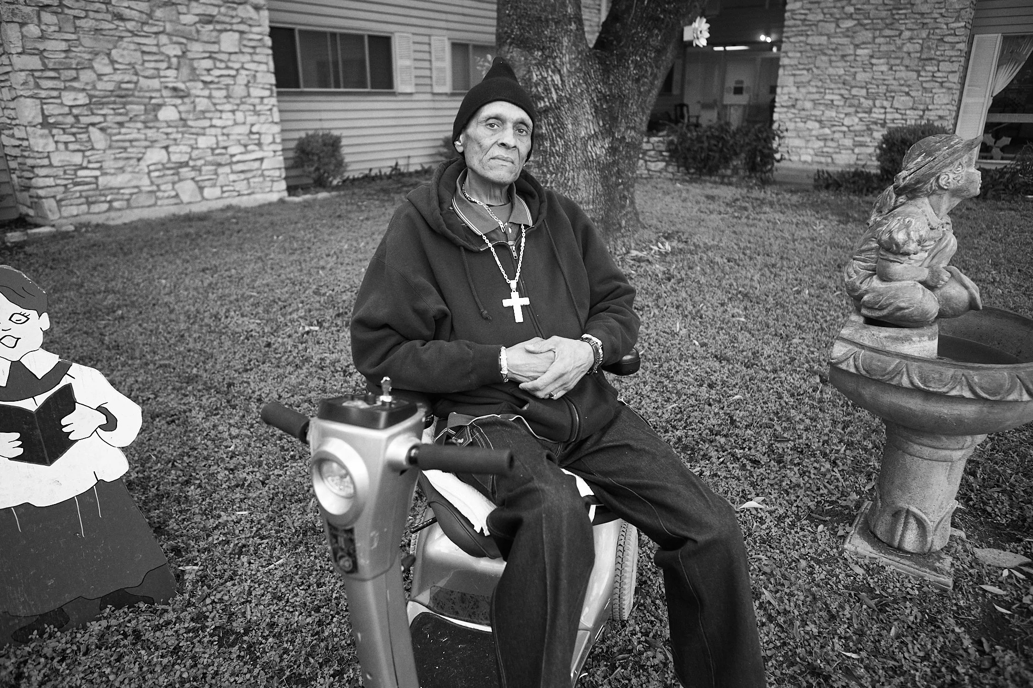 An elderly man seated on a mobility scooter in a garden, wearing a black hoodie, a black beanie, and a large cross necklace, with a serious expression. There is a cartoon cutout of a girl reading a book on his left, and a decorative garden statue of 