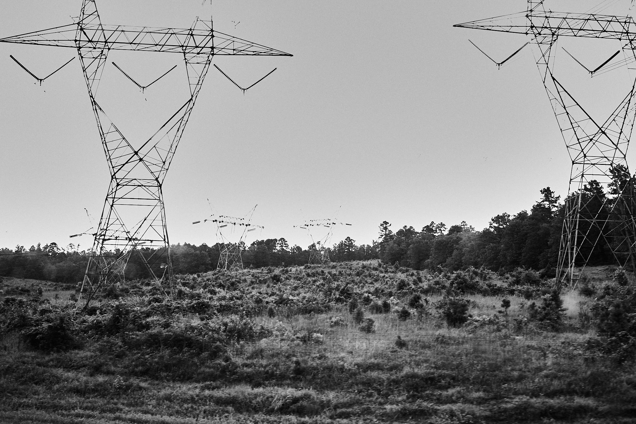 Black and white photo of electrical high-voltage power lines and towers in a rural landscape with trees and bushes.