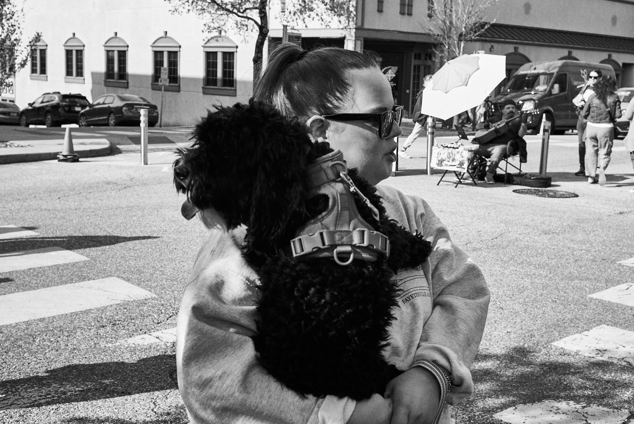 A woman carrying a dog on her shoulder in an outdoor urban setting with pedestrians, vehicles, and a street performer in the background.