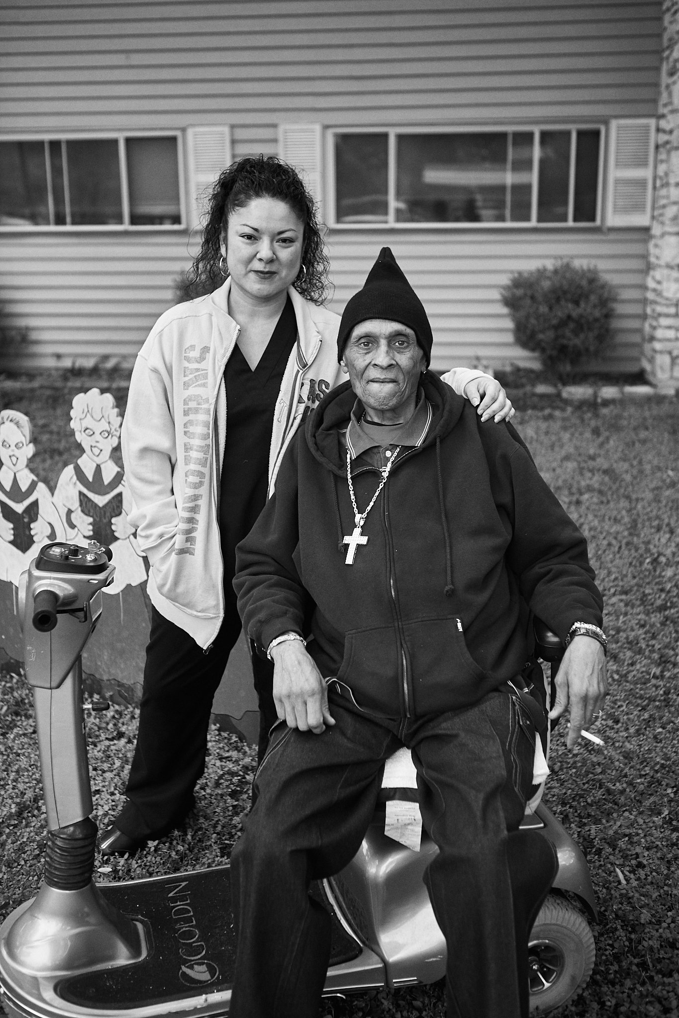 A woman standing beside a man sitting on a mobility scooter outdoors in front of a house. There are cartoon characters in the background and a scooter in the foreground.
