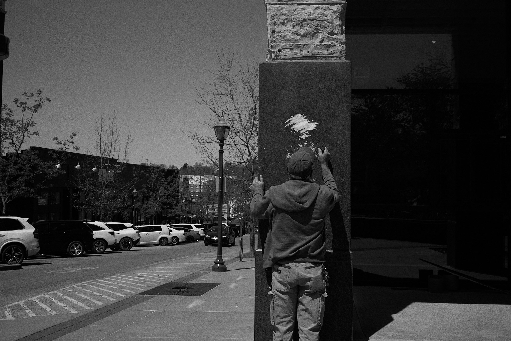 A person wearing a hoodie and cap creating street art on a wall with spray paint in an urban area with parked cars, trees, and buildings in the background.