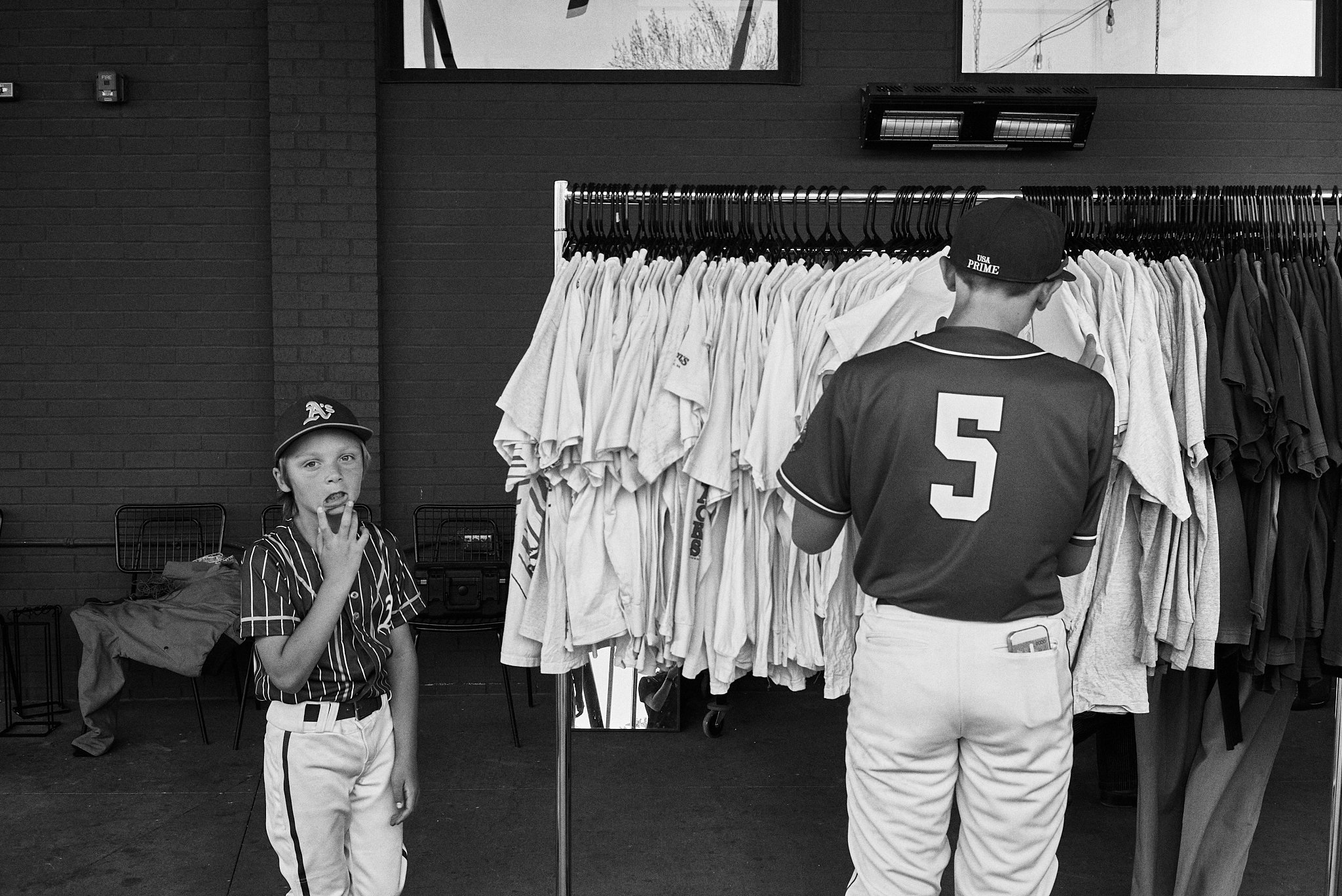A young boy in a baseball uniform eating a snack, standing in front of a clothing rack filled with various sports jerseys, as another person, seen from behind, browses the clothes.