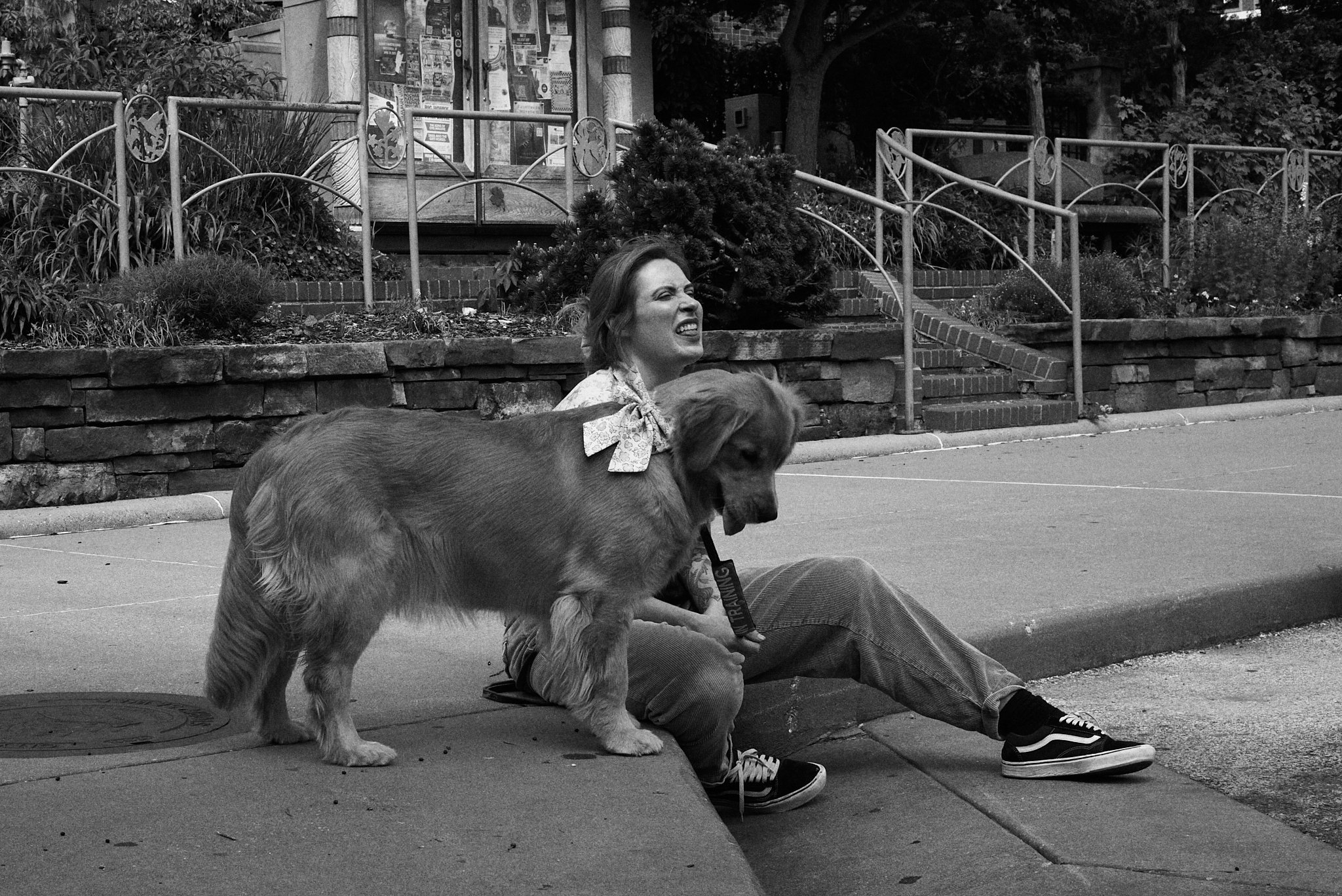A woman with short hair and casual clothing sitting on a curb, grimacing while a large dog with a bow on its collar stands close by.