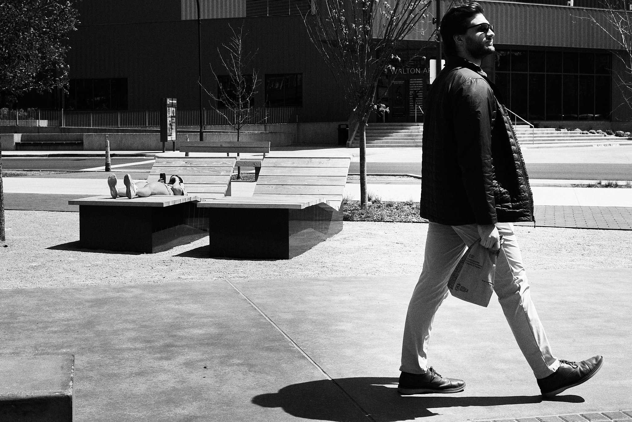 A man wearing glasses and a leather jacket walking on a sidewalk while carrying a paper bag, with a woman lying on a bench in the background in an urban park setting.