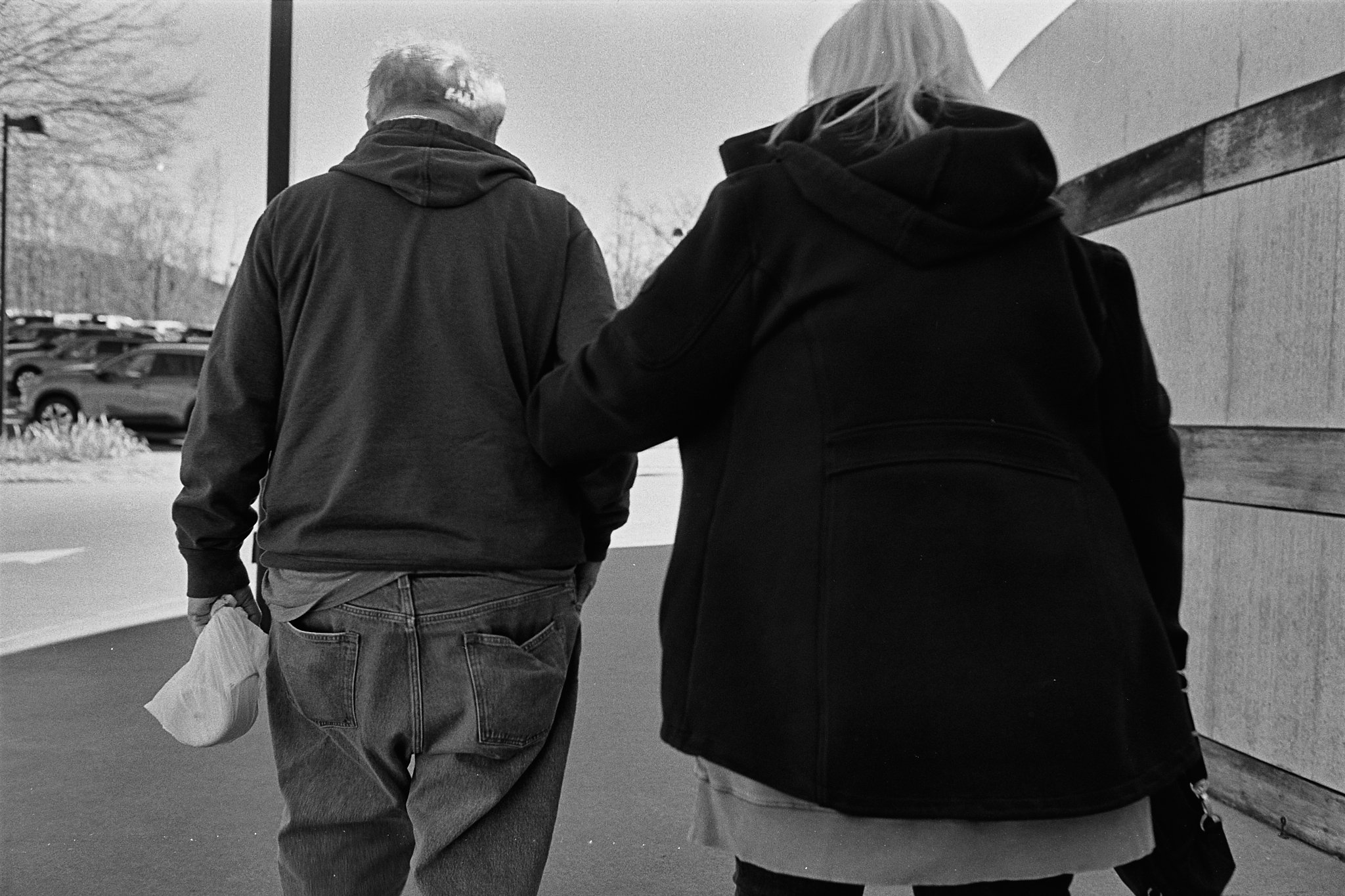Two people walking outdoors, one man and one woman, captured from behind, with parked cars and a building in the background.