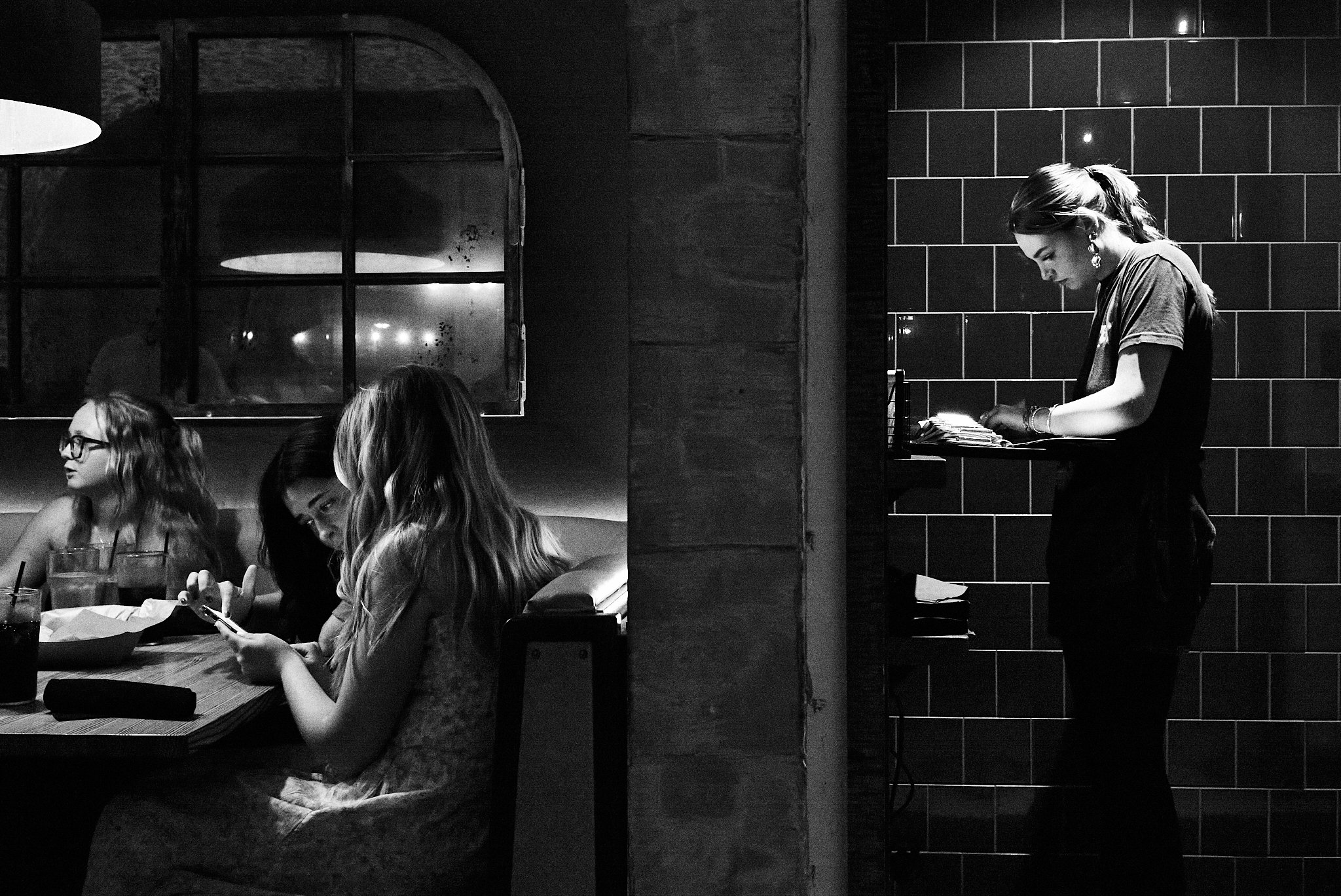 Black and white photo of women at a restaurant, three women sitting at a table on the left, and a waitress standing by the counter on the right.