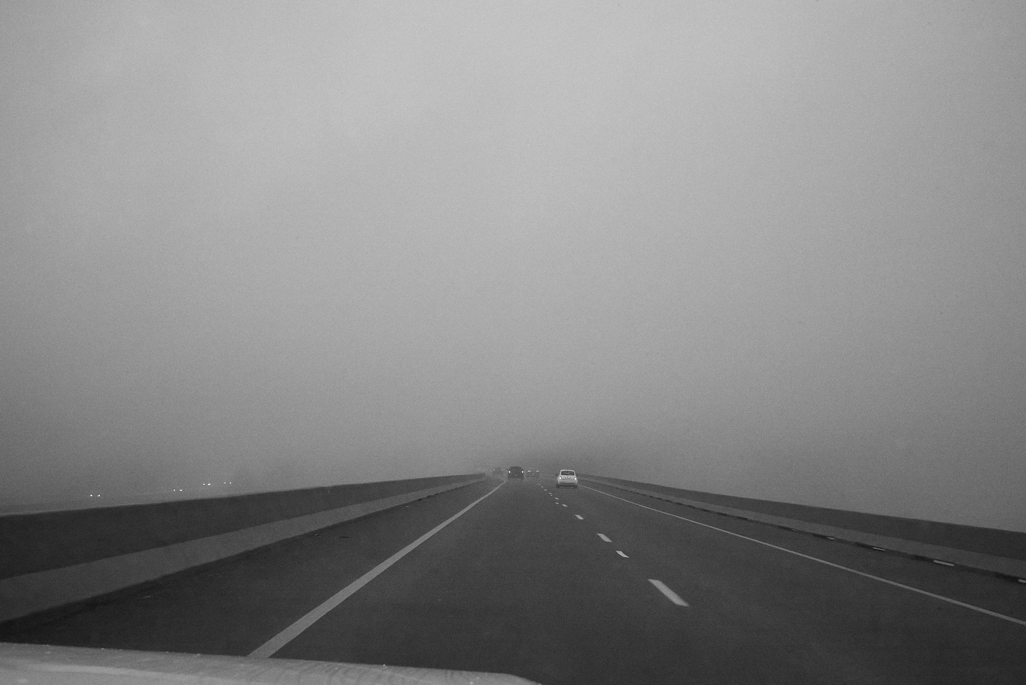 A black and white photo of a highway with several cars driving away in the distance, seen from inside a vehicle. The sky appears overcast or foggy.