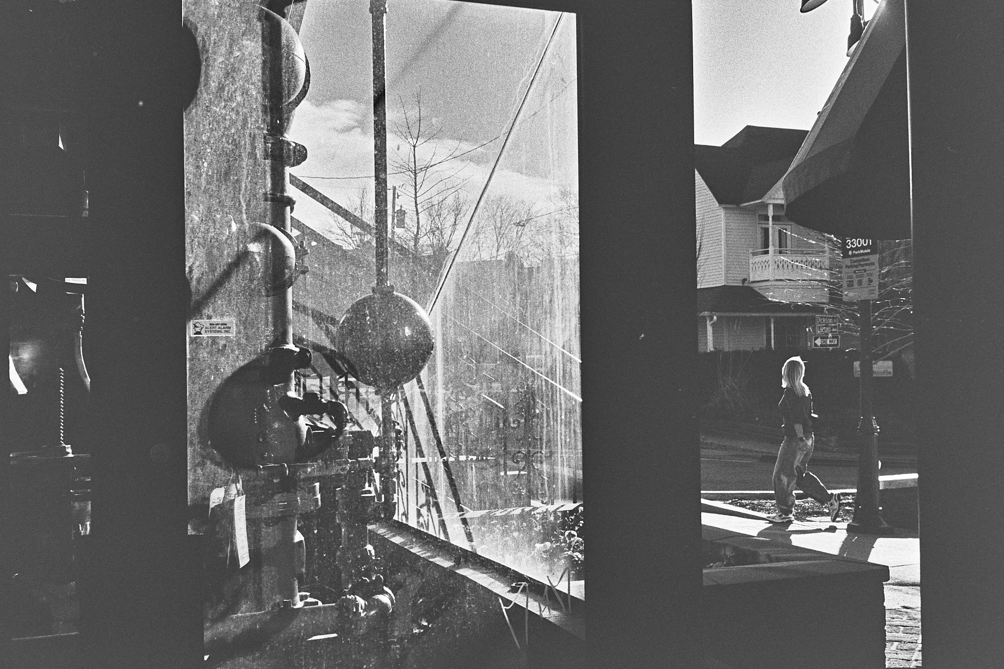 Black and white photo taken from inside a building, showing a large glass window with water droplets and a mechanical system with pipes on the left side. Outside, a person with long hair is walking on a sidewalk, illuminated by sunlight, with houses,