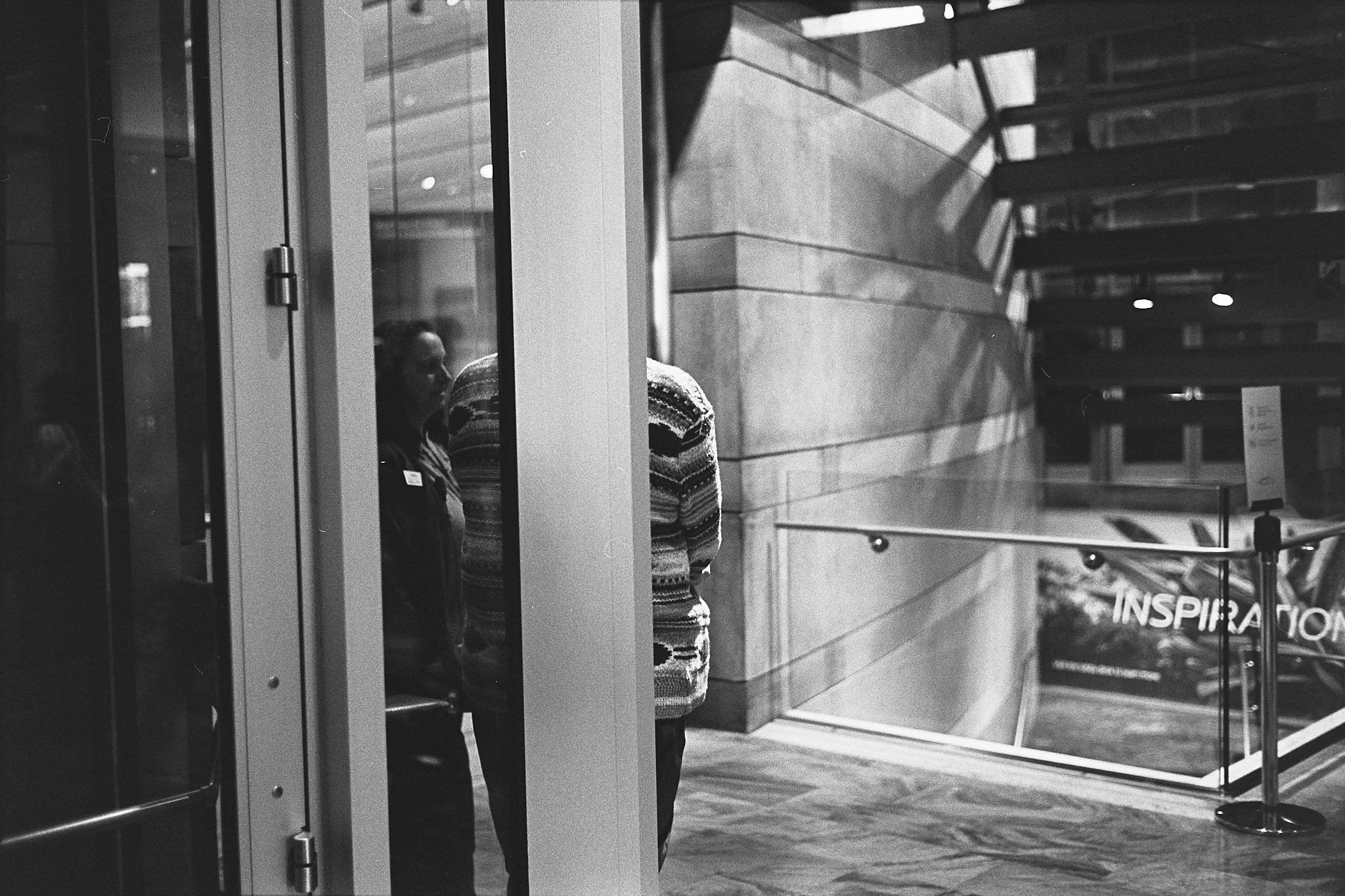 A woman standing underground next to a person with their back to the camera in a modern interior space with stairs, glass walls, and a railing.