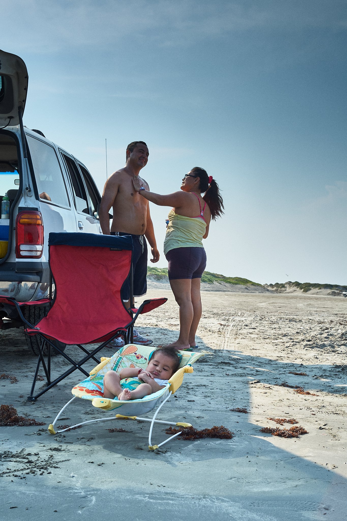 A family at the beach shows a woman applying sunscreen to a shirtless man while a sleeping baby lies in a yellow baby bouncer on the sand. Beach gear is near an open vehicle, and the sky is clear with a distant shoreline.