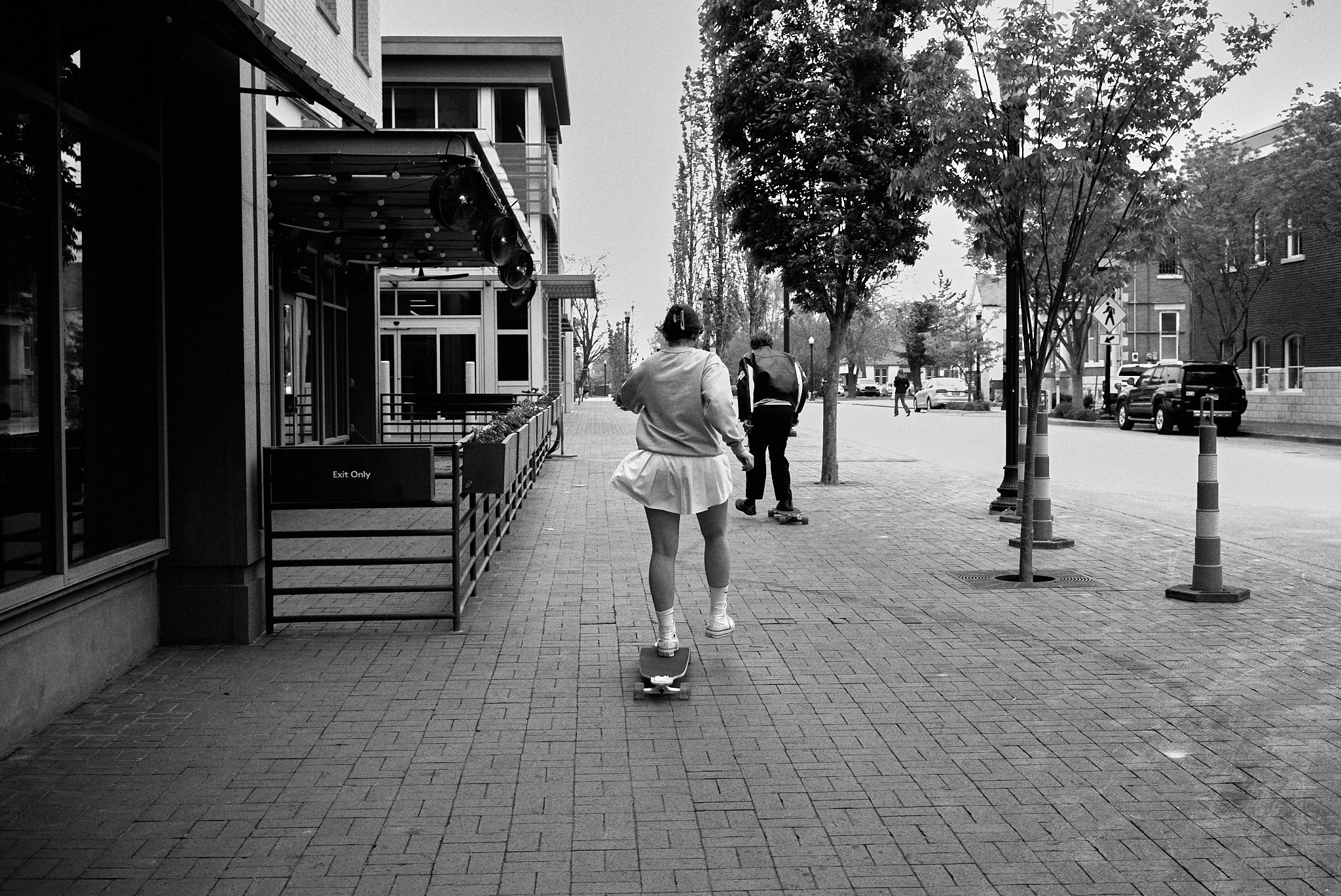 A person skateboarding on a city sidewalk with trees, buildings, and parked cars in the background, black and white photo.