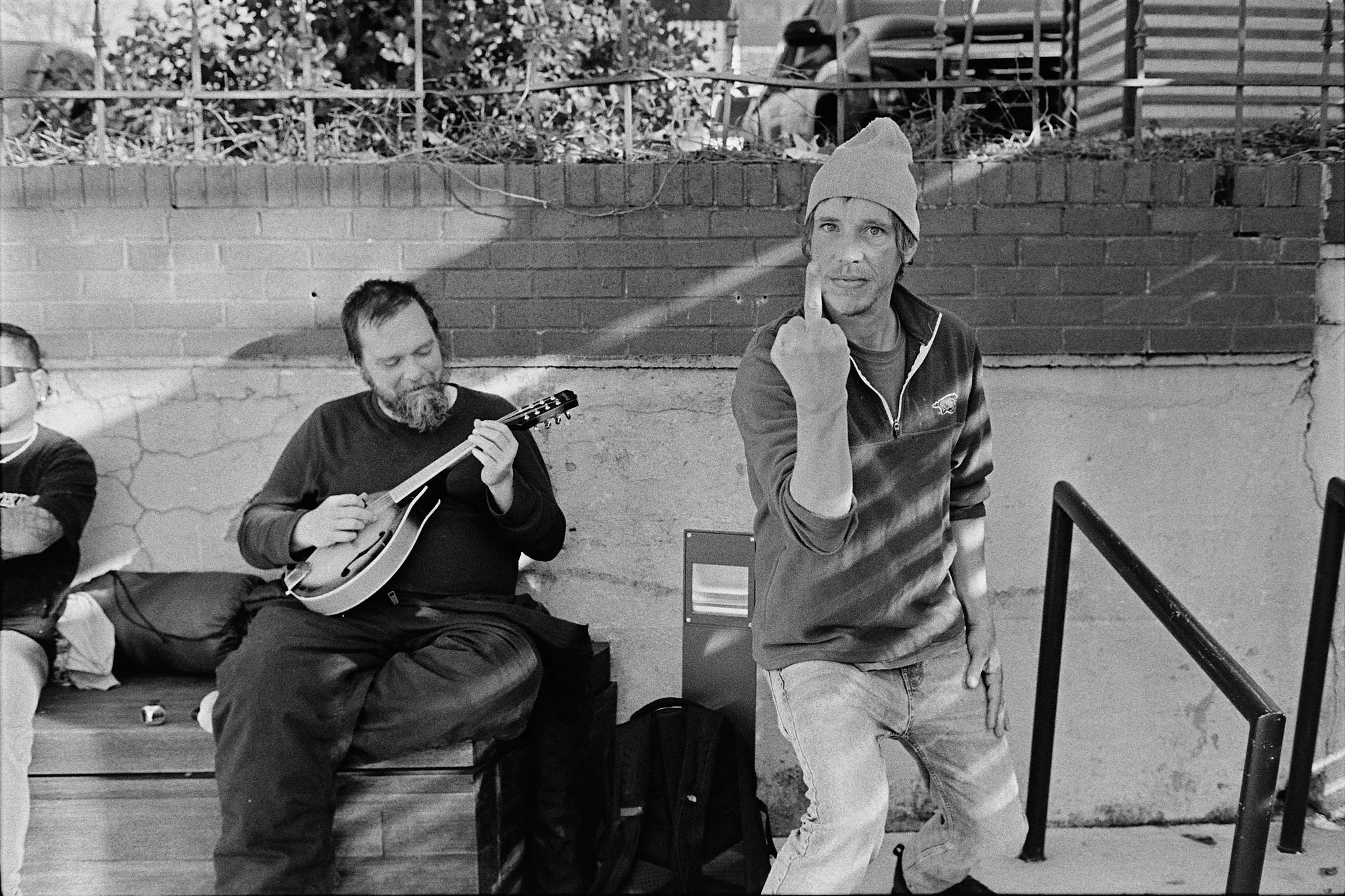 A street scene in black and white with two men. One man is sitting on a bench playing a guitar, wearing a dark long-sleeve shirt with a beard. The other man, wearing a beanie and a hoodie, is standing with one hand on his thigh and the other hand rai