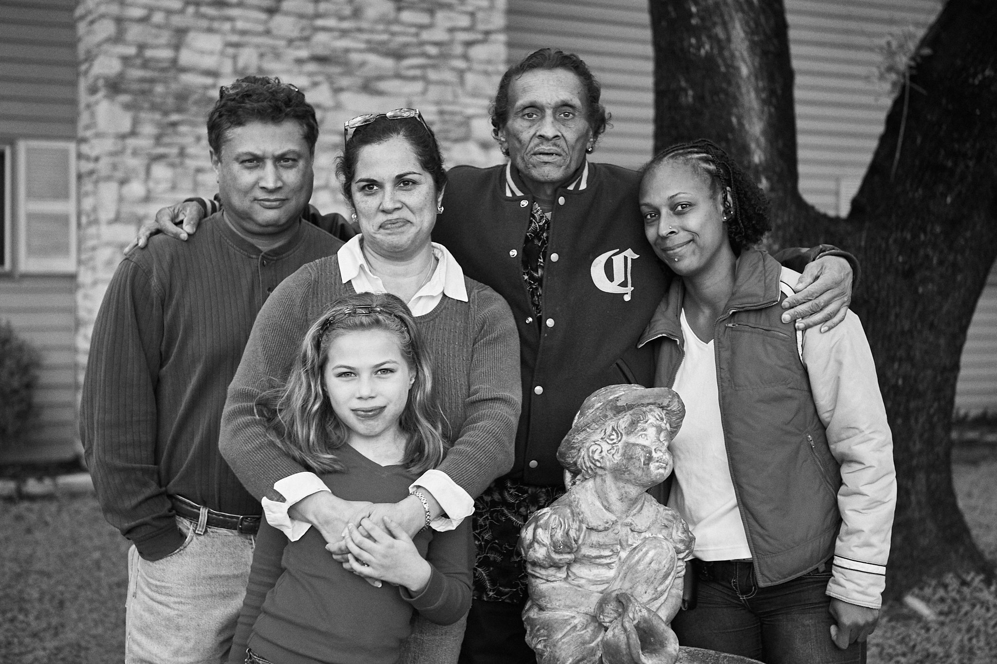 Group of five people, three women and two men, posing outdoors in front of a tree and a house, with a small statue of a woman holding a corn cob in front of them, in black and white.