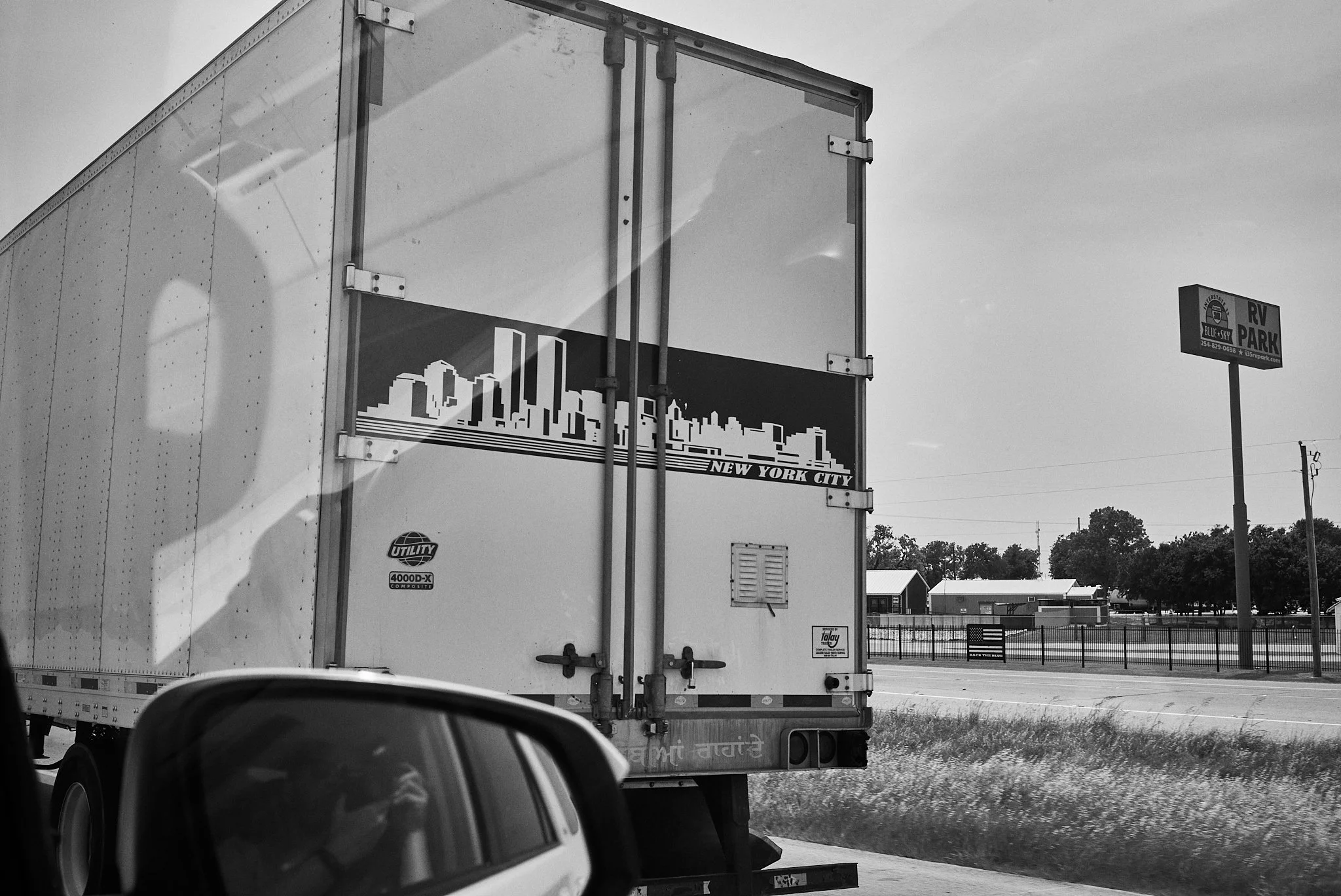 A large truck on the highway with a New York City skyline graphic on the back, next to a sign reading "RV Park." The image is in black and white.