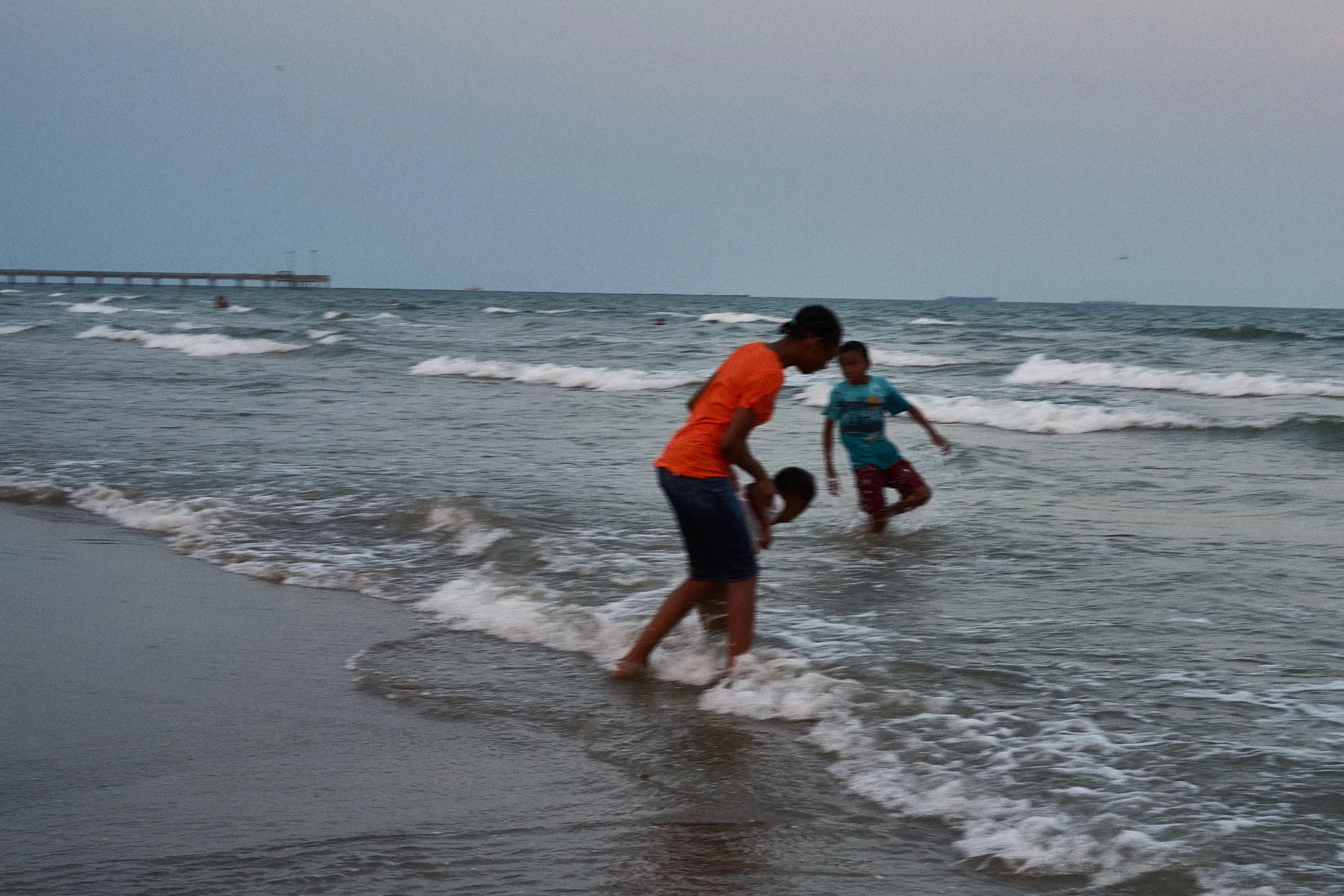 Two children playing in the ocean with a pier visible in the distance, waves coming onto the sandy shore during dusk.