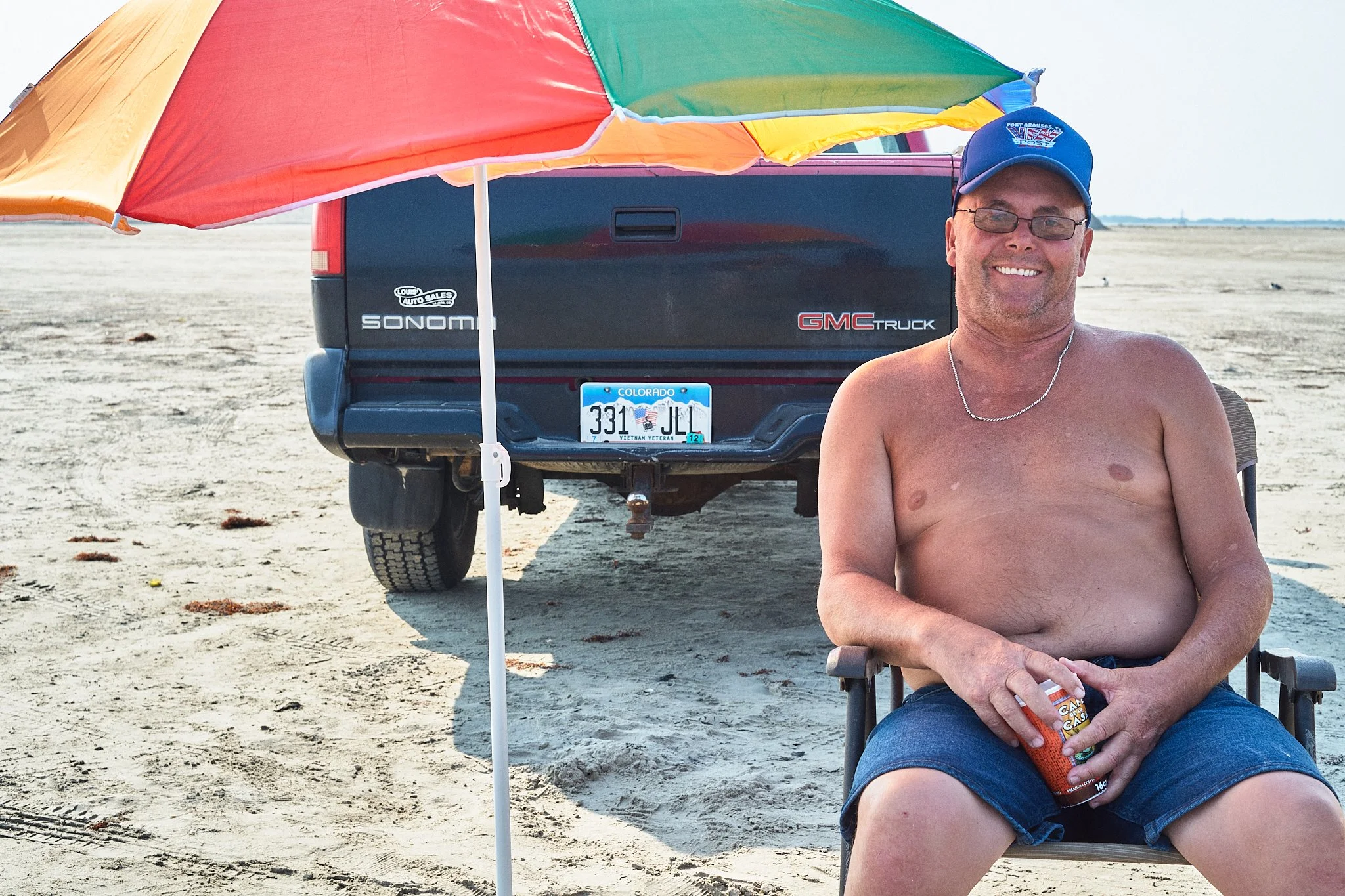 Smiling man sitting under a multicolored umbrella on a beach, holding a can of beer, with a black GMC truck and Colorado license plate in the background.