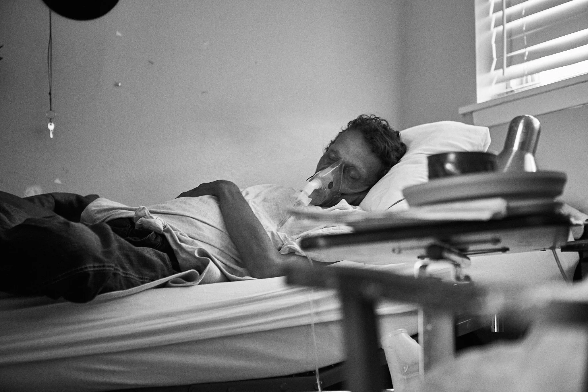 A person lying in a hospital bed with an oxygen mask, appearing to be asleep or unconscious, next to a window with blinds.