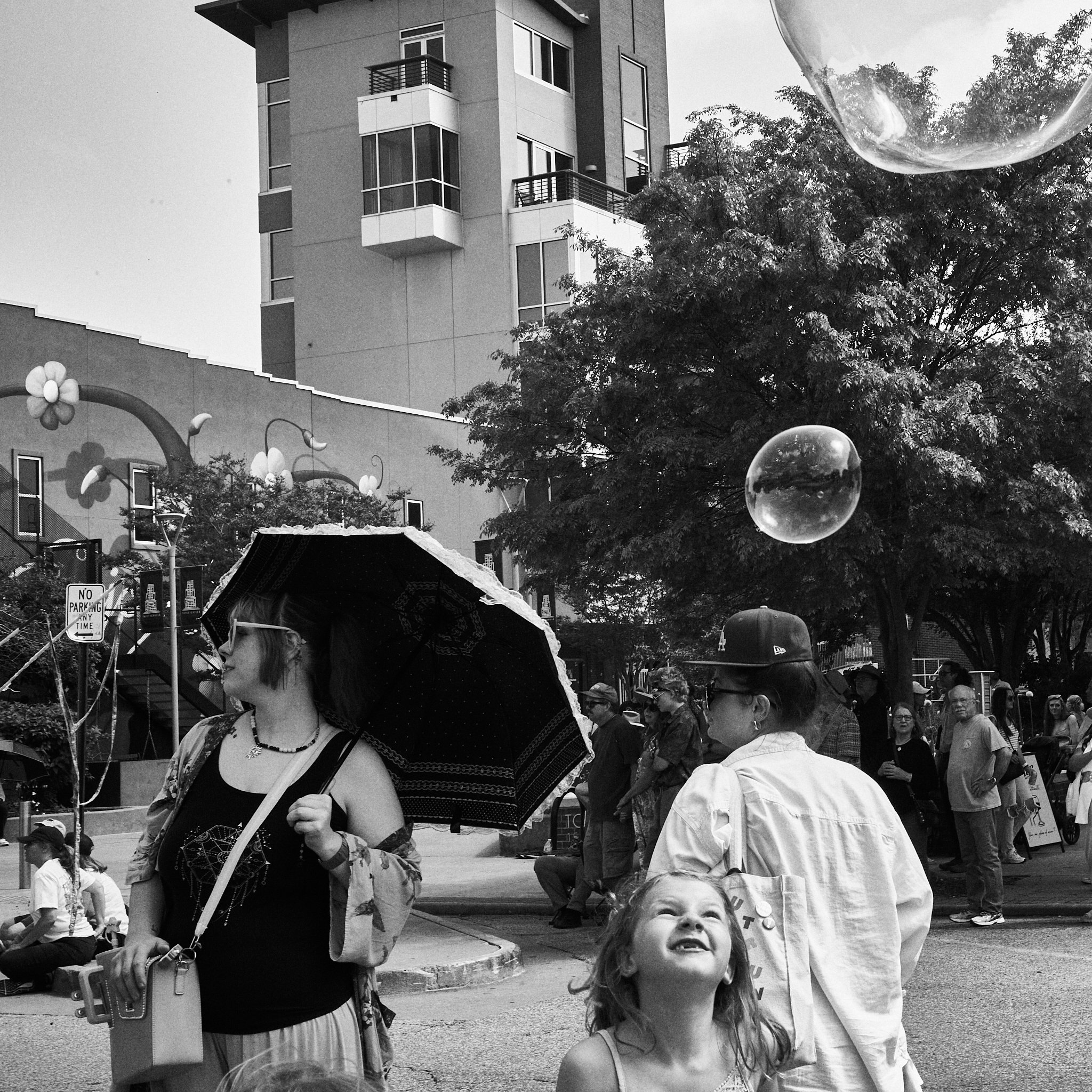 People gathered outdoors in a city street under a tree, with some holding umbrellas, balloons, and cameras; a building with balconies is in the background.