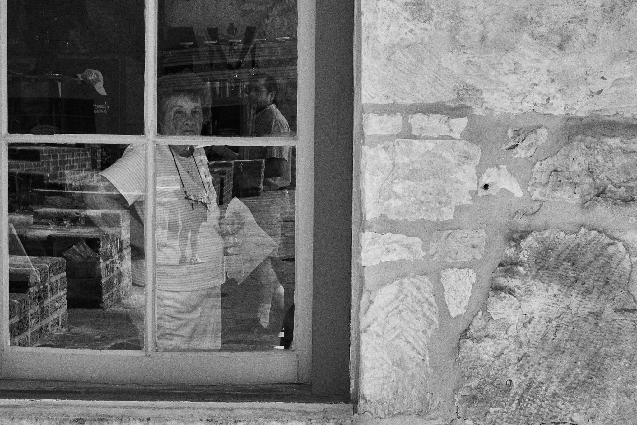 Reflected image of an elderly woman with short hair and glasses hanging around her neck, visible through a window with a wooden frame. She is inside a room with bookshelves and brick walls, and appears to be looking at the camera.