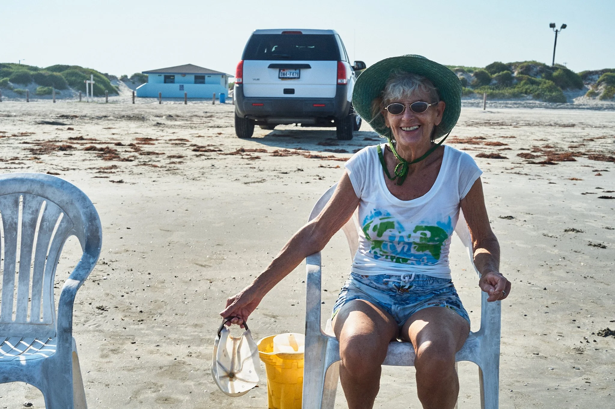 An elderly woman with gray hair, wearing sunglasses, a wide-brimmed green hat, a white T-shirt with a colorful design, and blue patterned shorts, sits on a white plastic chair on a sandy beach. She has a broad smile, and is holding a white sun hat in