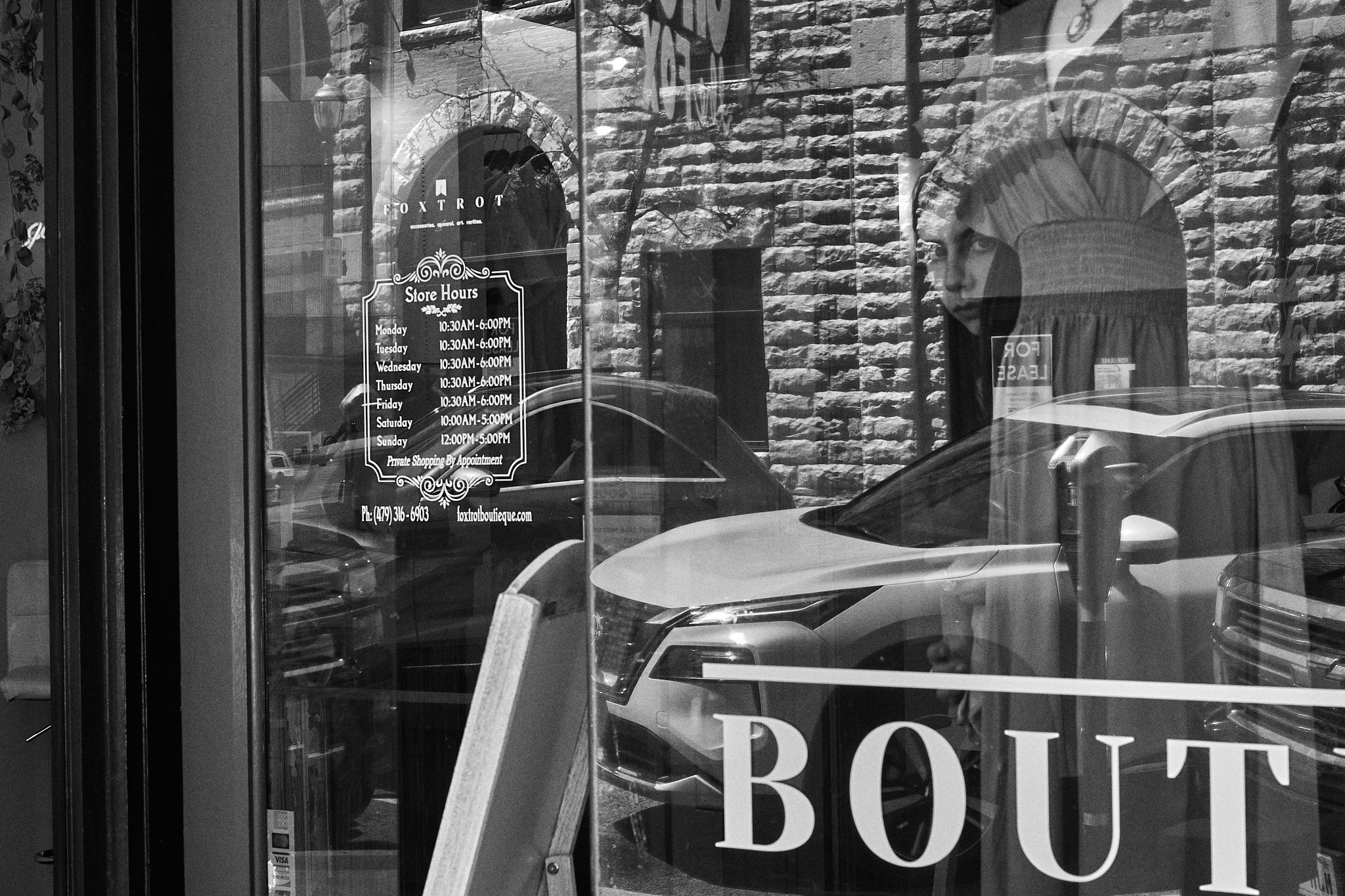 Reflective black-and-white photo of a storefront window with signage displaying store hours and a glimpse of a person's face reflected in the glass, along with parked cars outside and brick buildings in the background.