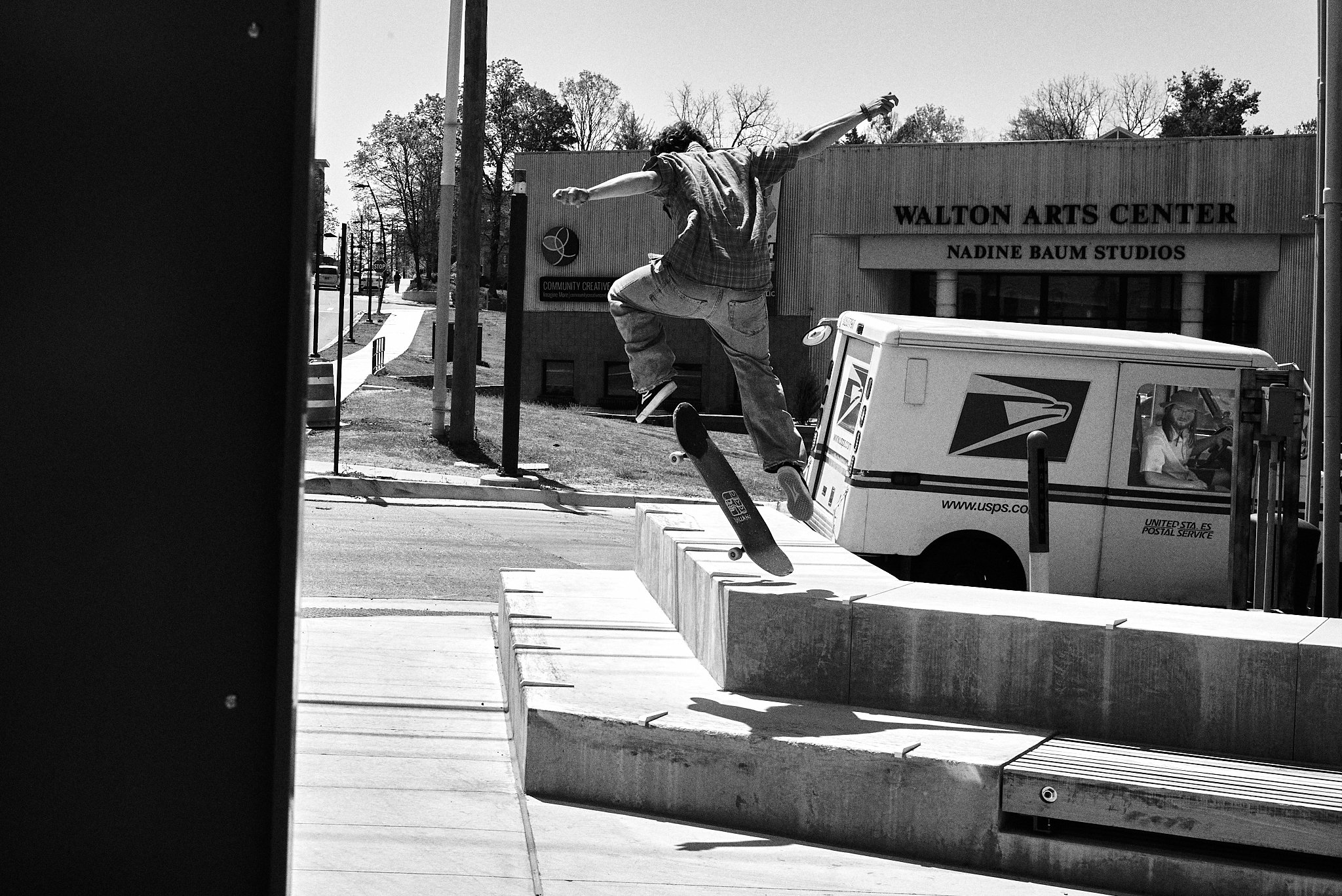 A skateboarder performing a trick on a ledge in front of a USPS mail truck, with a building labeled Walton Arts Center in the background.