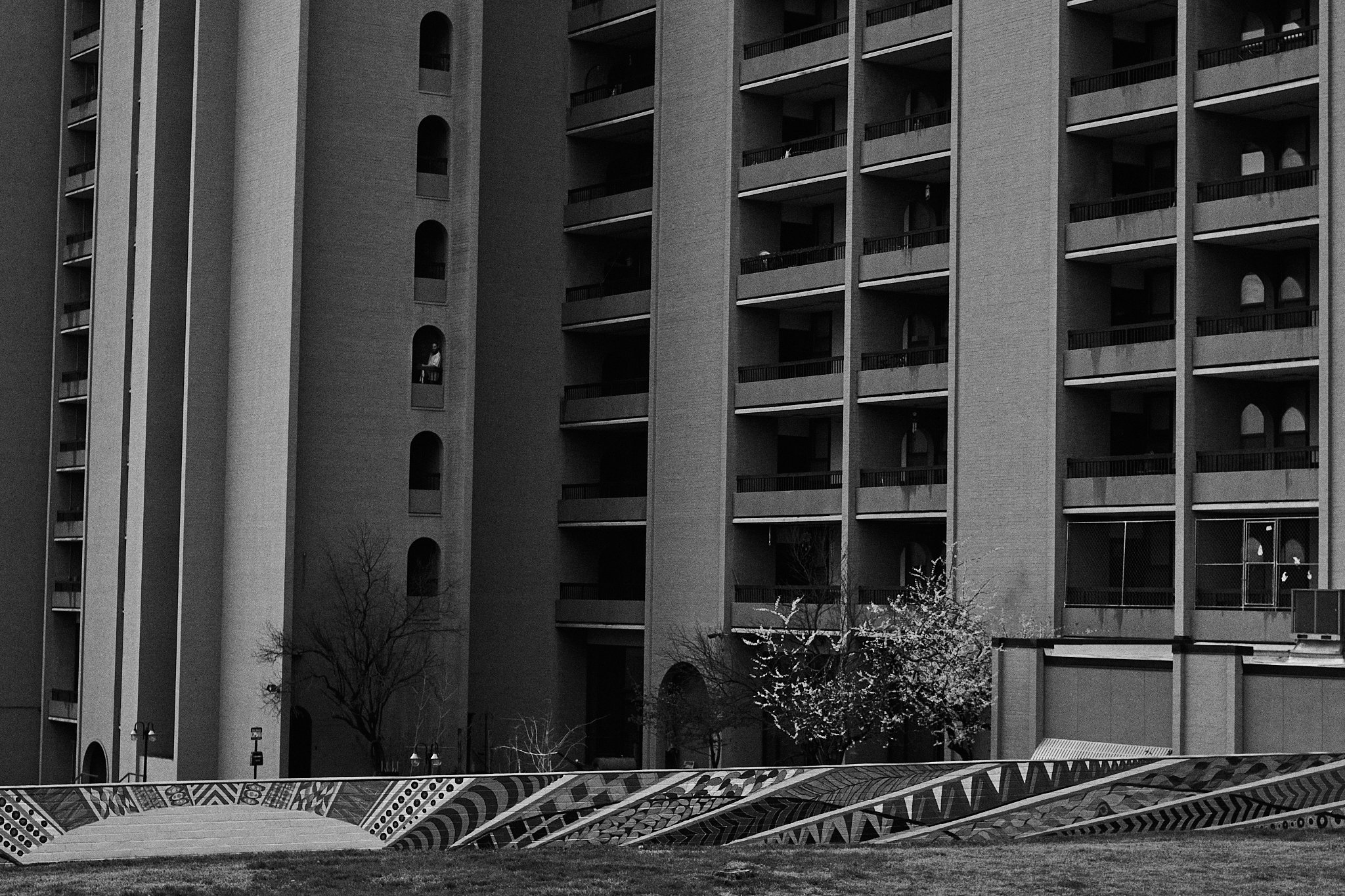 Black and white photo of a large multi-story apartment building with balconies. There are some trees and a decorative mural in the foreground.