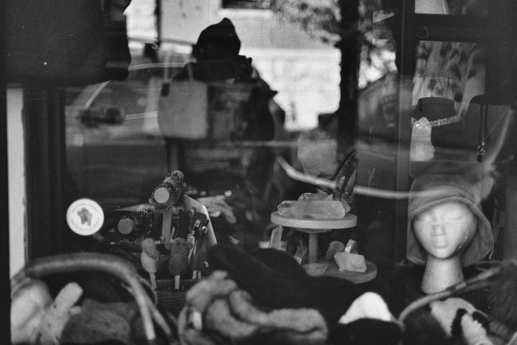View through a store window showing various toys including a mannequin head with a wig, small figurines, and other miscellaneous items, with a person's reflection in the glass.