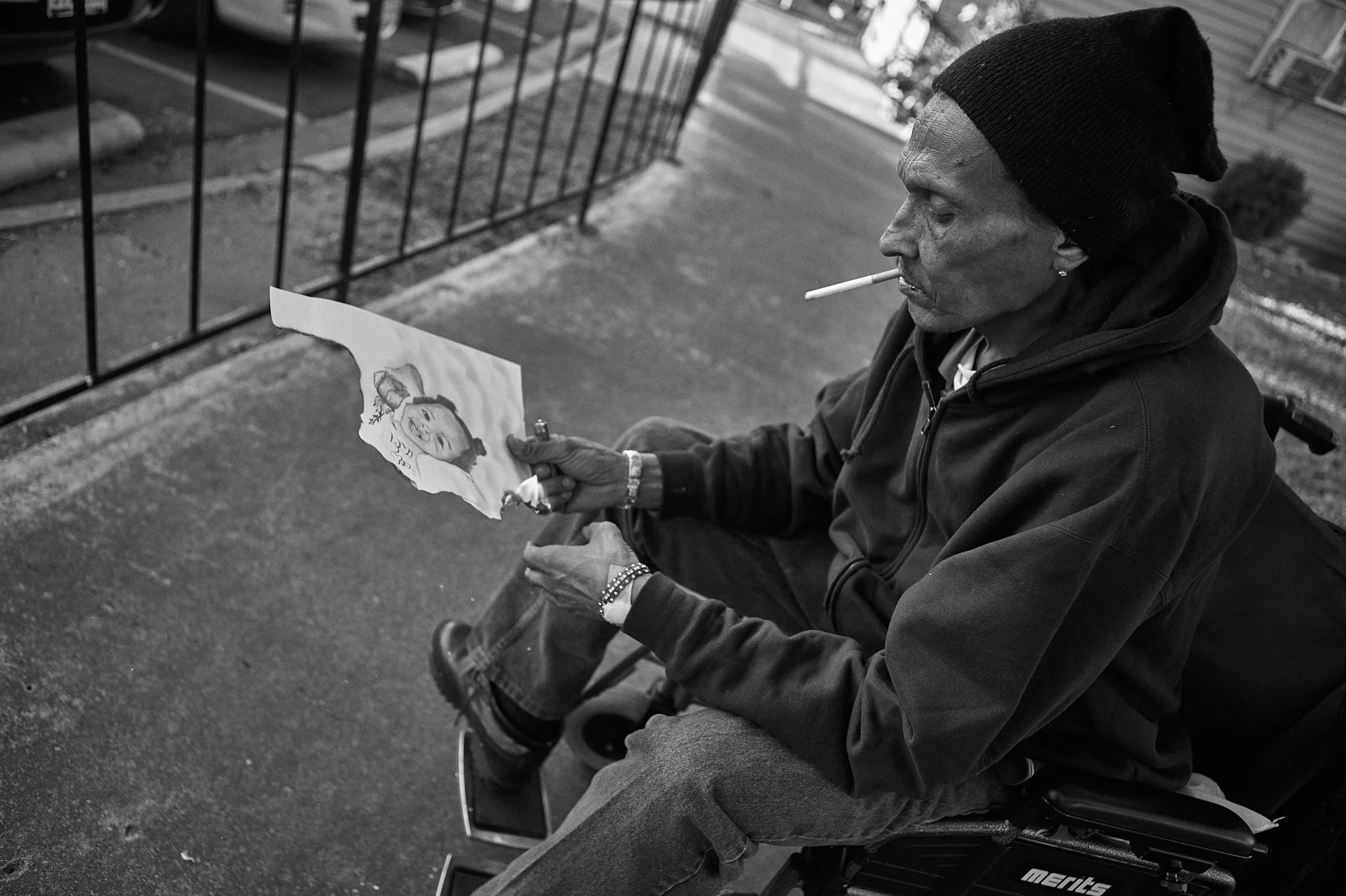A man sitting in a wheelchair outdoors, smoking a cigarette, and looking at a torn photograph of a woman.