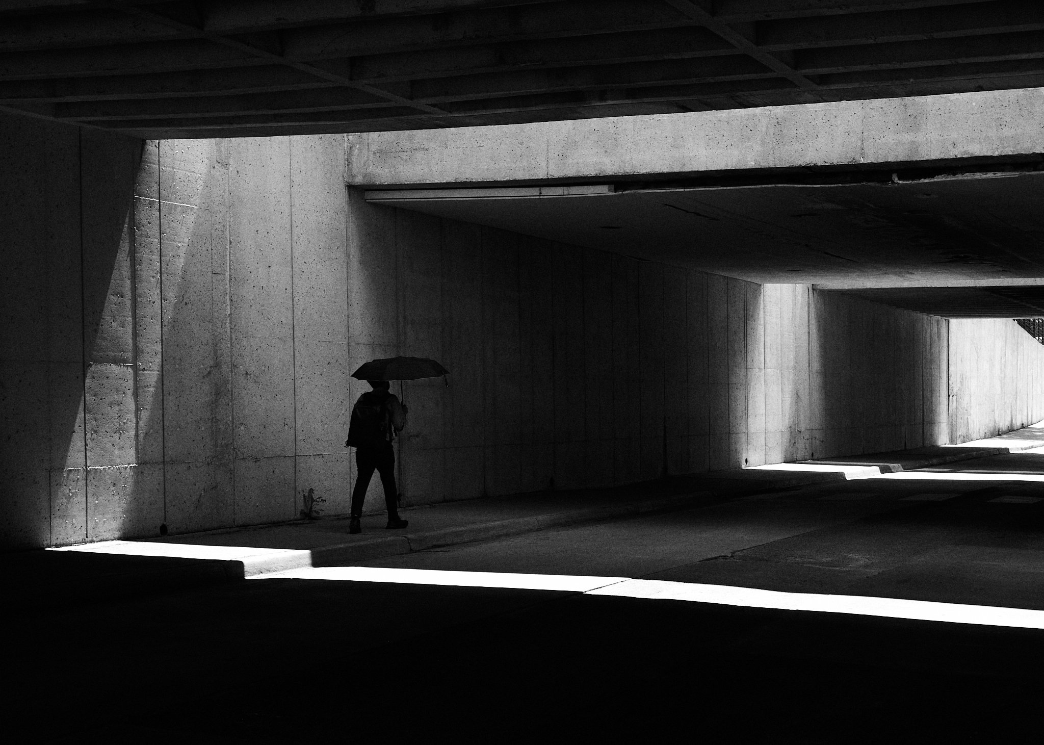 A person walking with an umbrella in an underpass with shadows and concrete walls.
