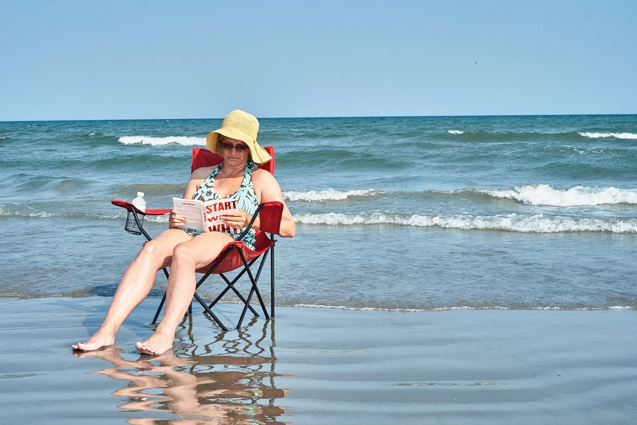 A woman sitting on a red beach chair at the beach, wearing a straw hat, sunglasses, and a floral swimsuit, reading a magazine with the ocean and small waves in the background.