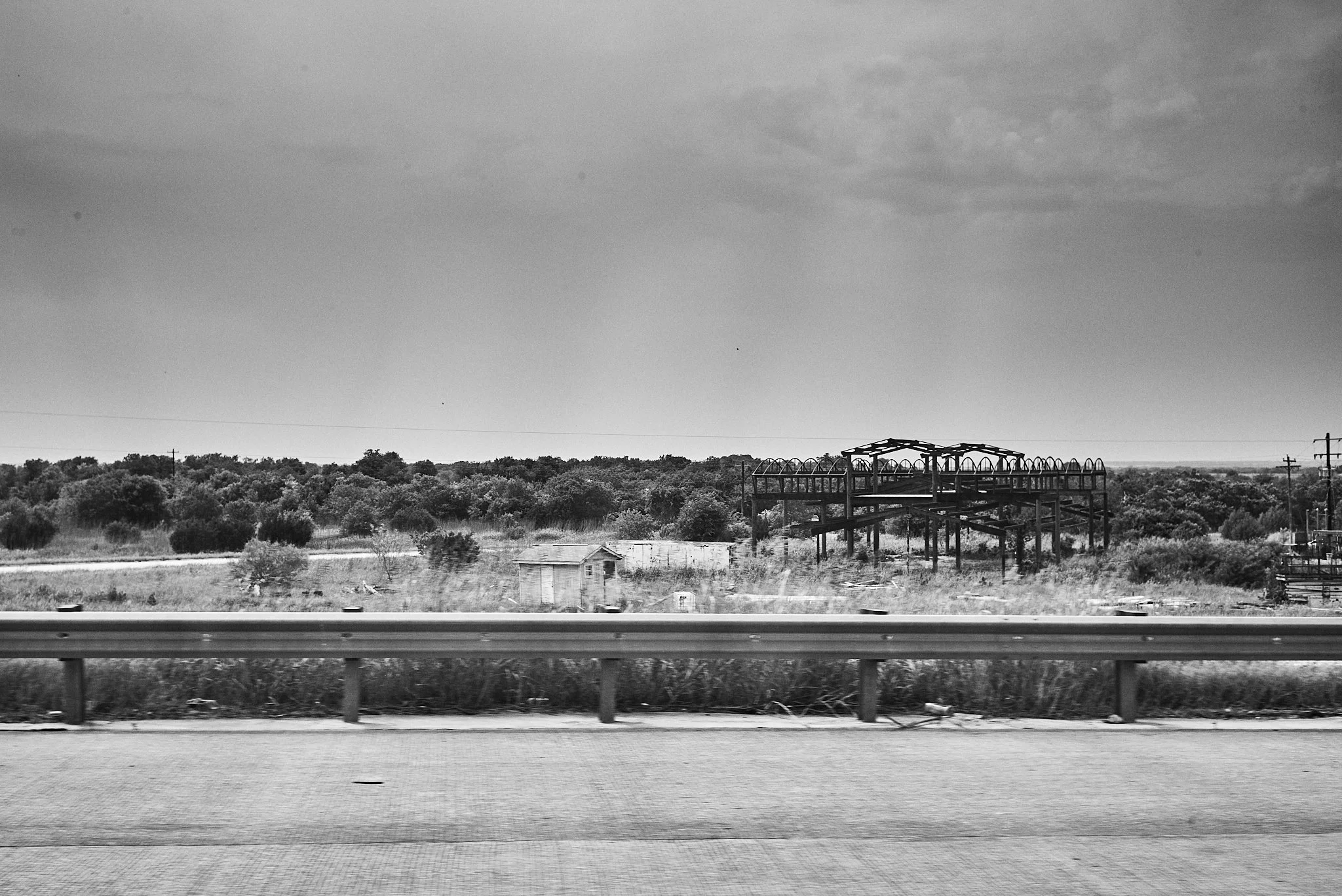 A black and white photo taken from a moving vehicle shows a rural landscape with overgrown grass and bushes, a small shed, and a large metallic structure, possibly a bridge or framework, in the distance under a cloudy sky.