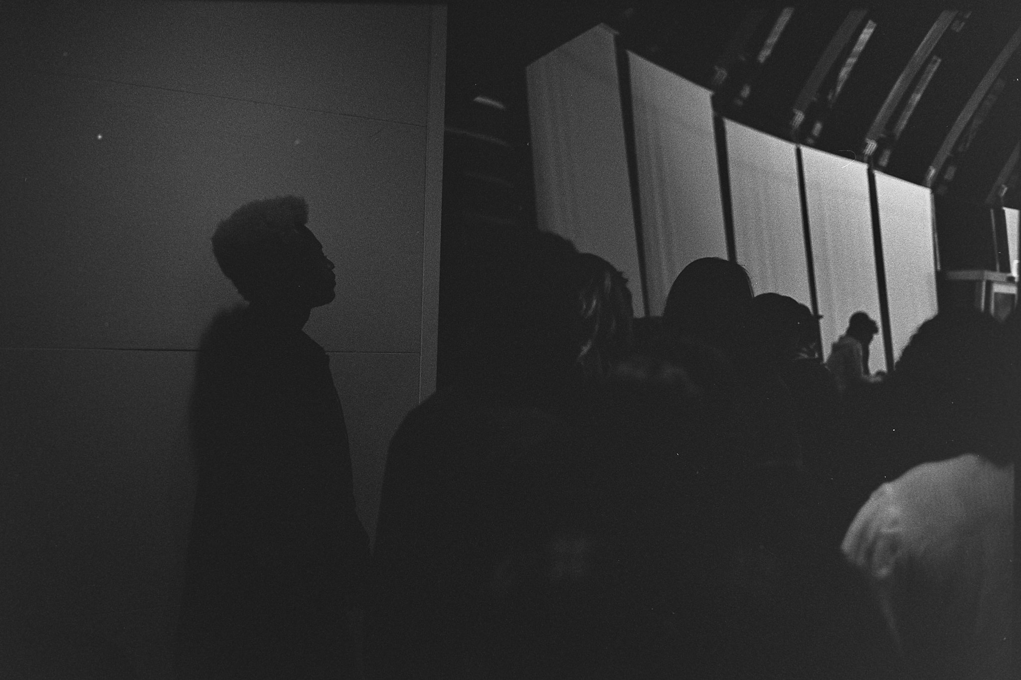 Silhouette of several people standing in a dark room with a large, illuminated wall or screen in the background.