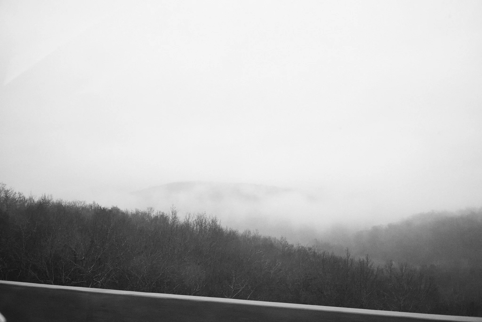 Black-and-white photo of a foggy landscape with bare trees and distant hills, viewed from a moving vehicle.