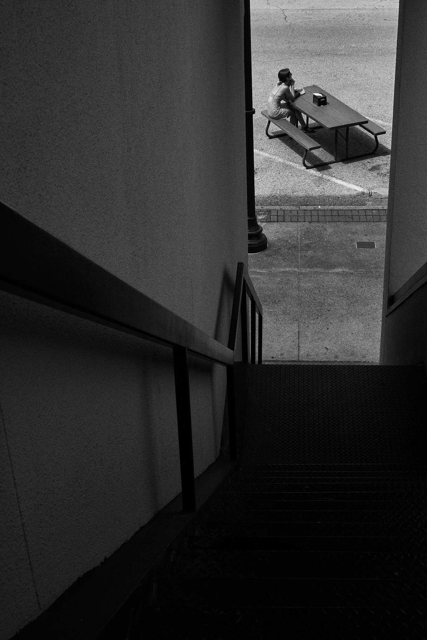 A person sitting alone at a picnic table outside, viewed from the top of a staircase in a black and white photo.