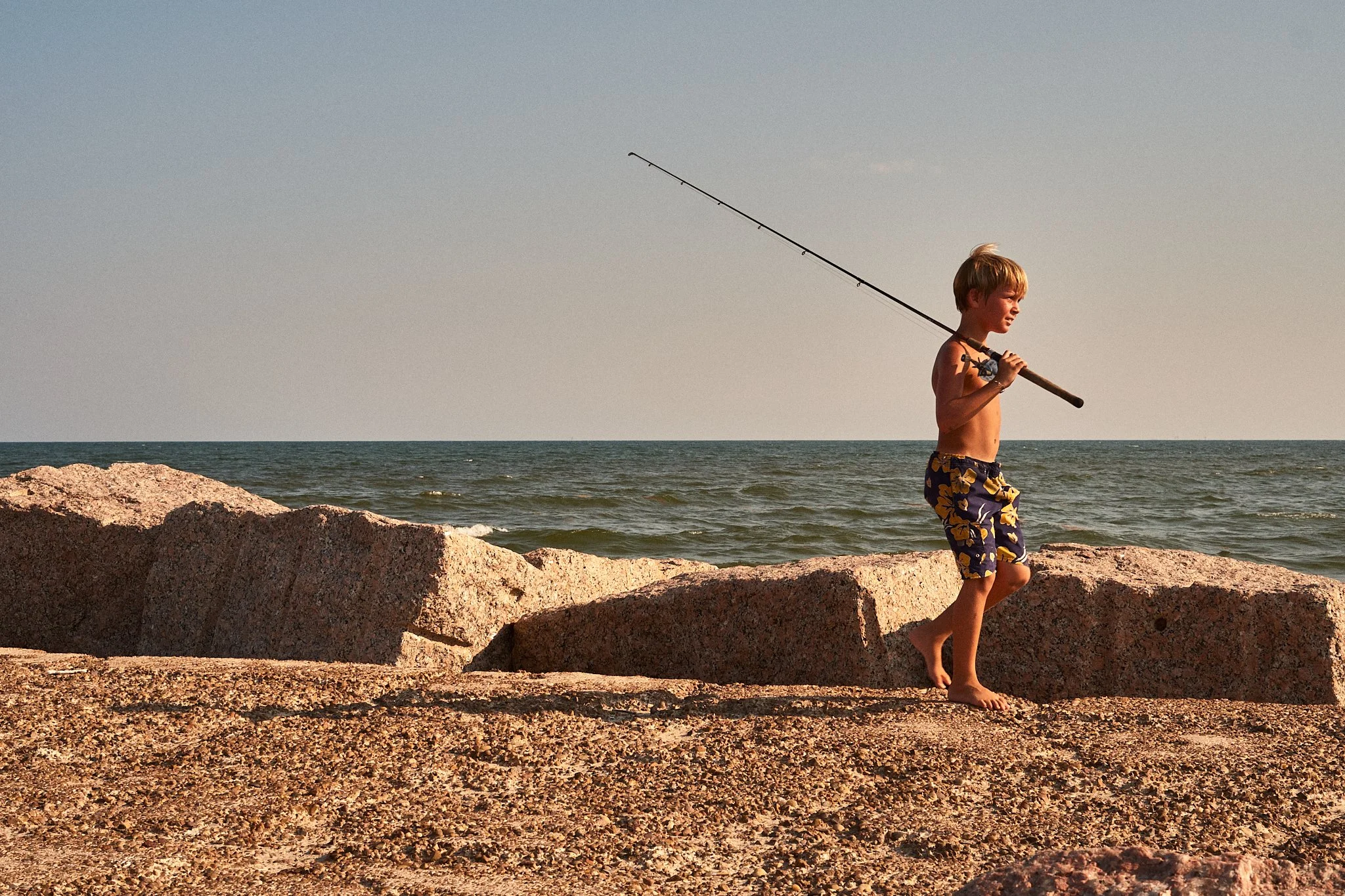 Young boy in swim trunks carrying a fishing rod walking on rocks along the beach with ocean waves in the background during sunset.