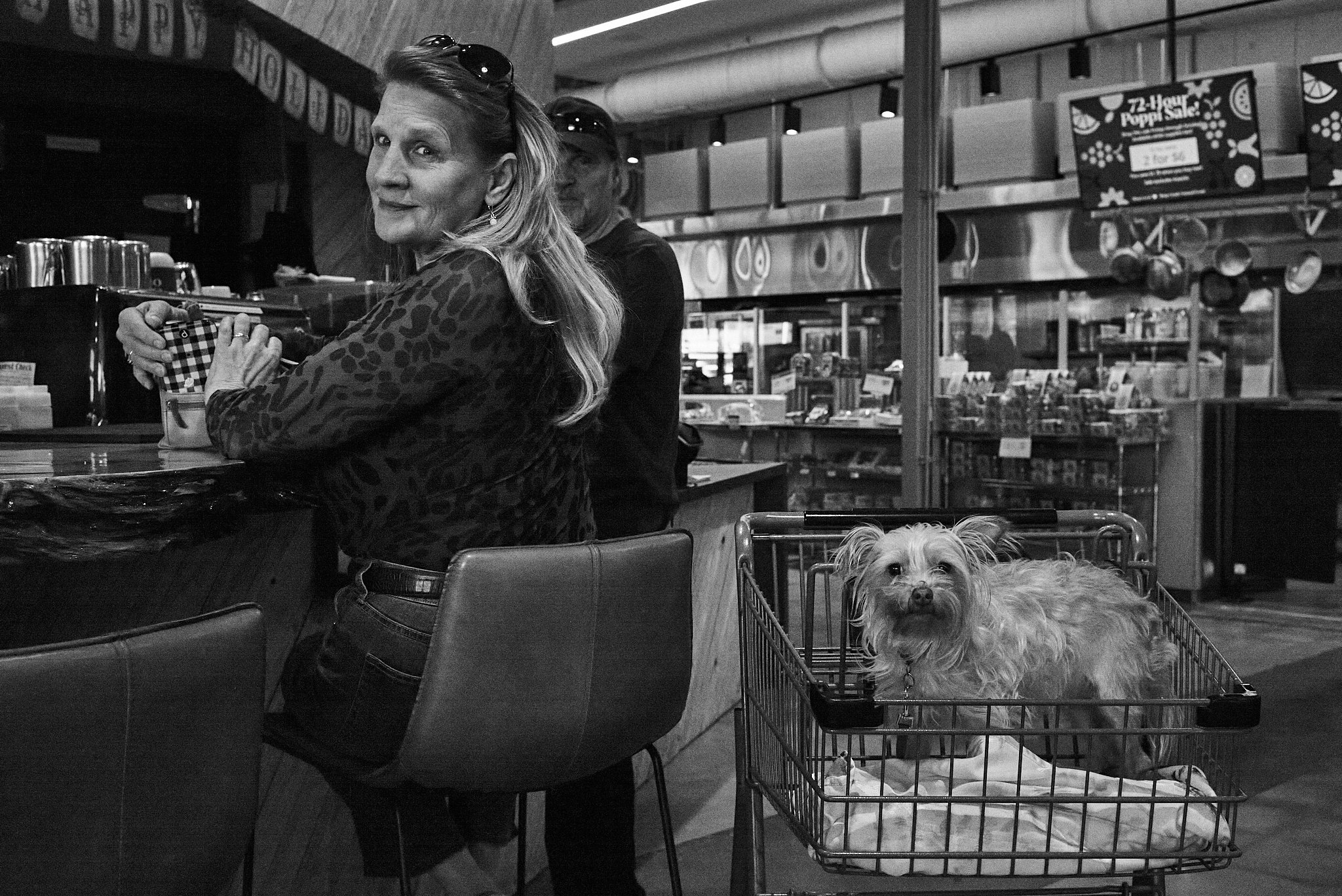 A woman leaning on a counter in a store or café, with a small dog in a shopping cart nearby, looking at the camera. The store has shelves with products and signs, and there are other people in the background.