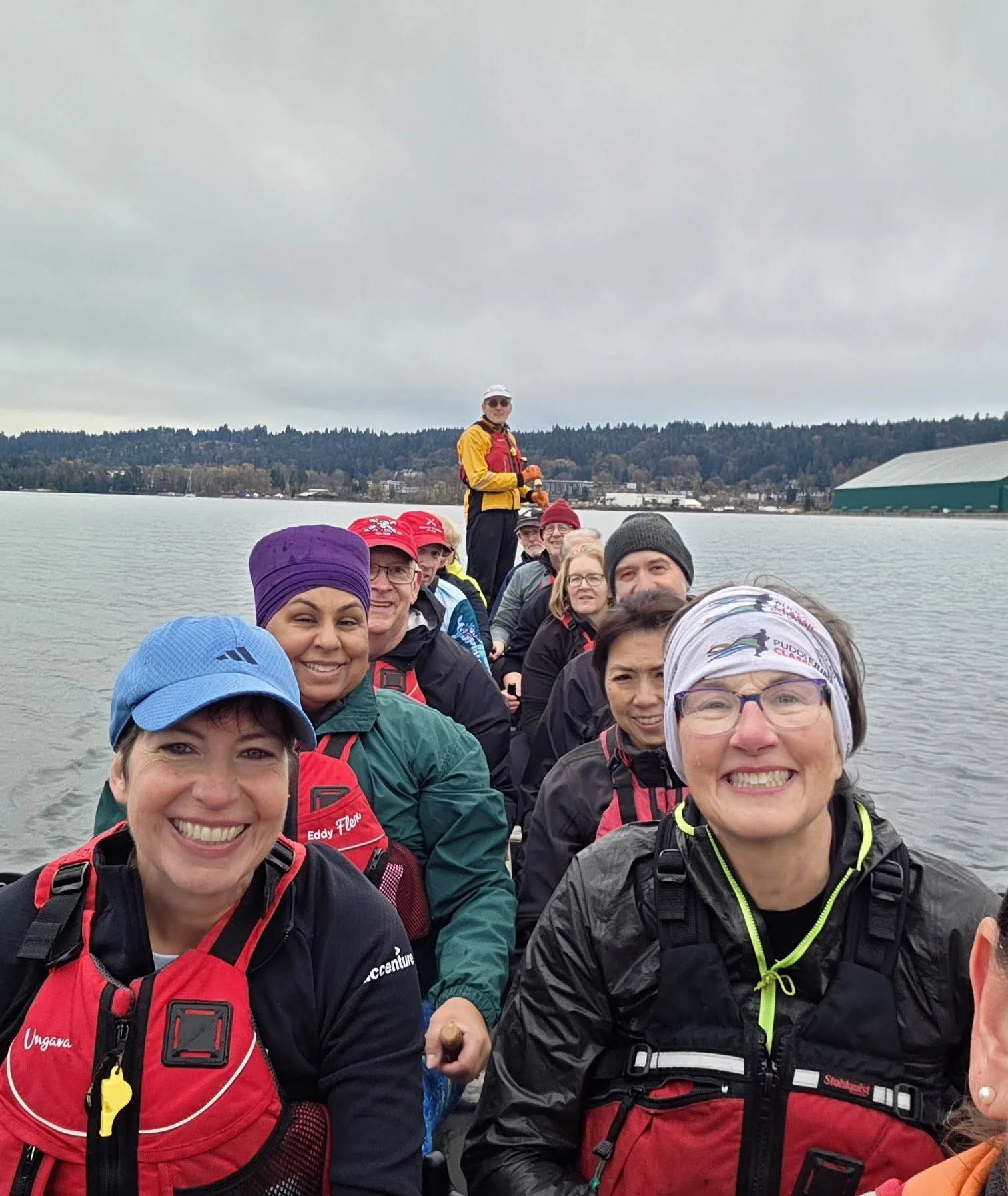 A group of people in outdoor gear smiling on a boat, with water and trees in the background.