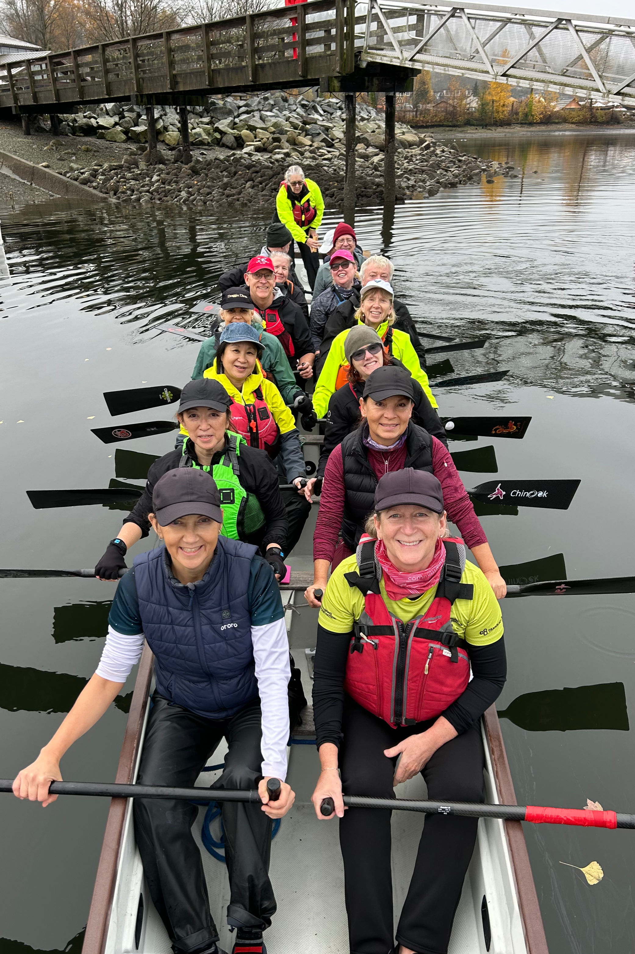 A group of people in a dragon boat for paddling on a calm river, with trees and a bridge in the background.