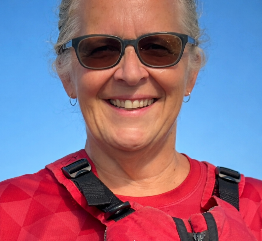 Close-up of an older woman smiling, wearing sunglasses, a red athletic shirt, and a harness against a clear blue sky.