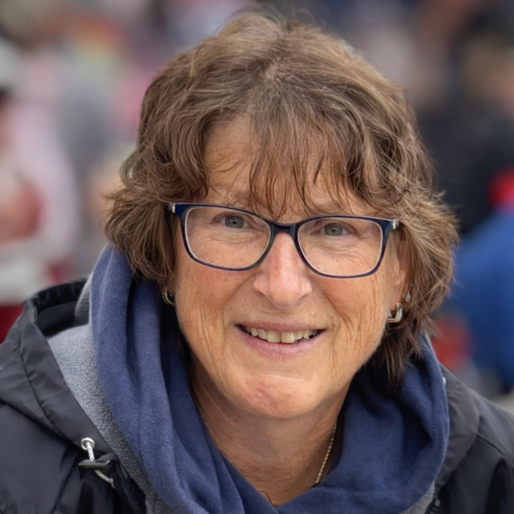 A middle-aged woman with short, wavy brown hair and glasses, smiling outdoors with a blurred crowd in the background.
