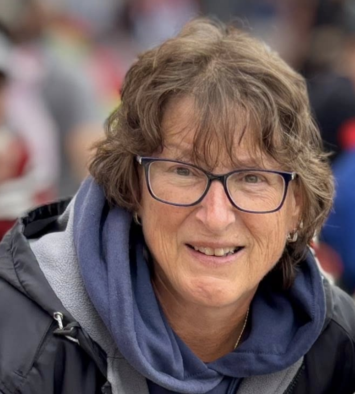 A smiling woman with glasses and short wavy brown hair, wearing a navy blue hoodie and a black jacket, outdoors in a crowd.
